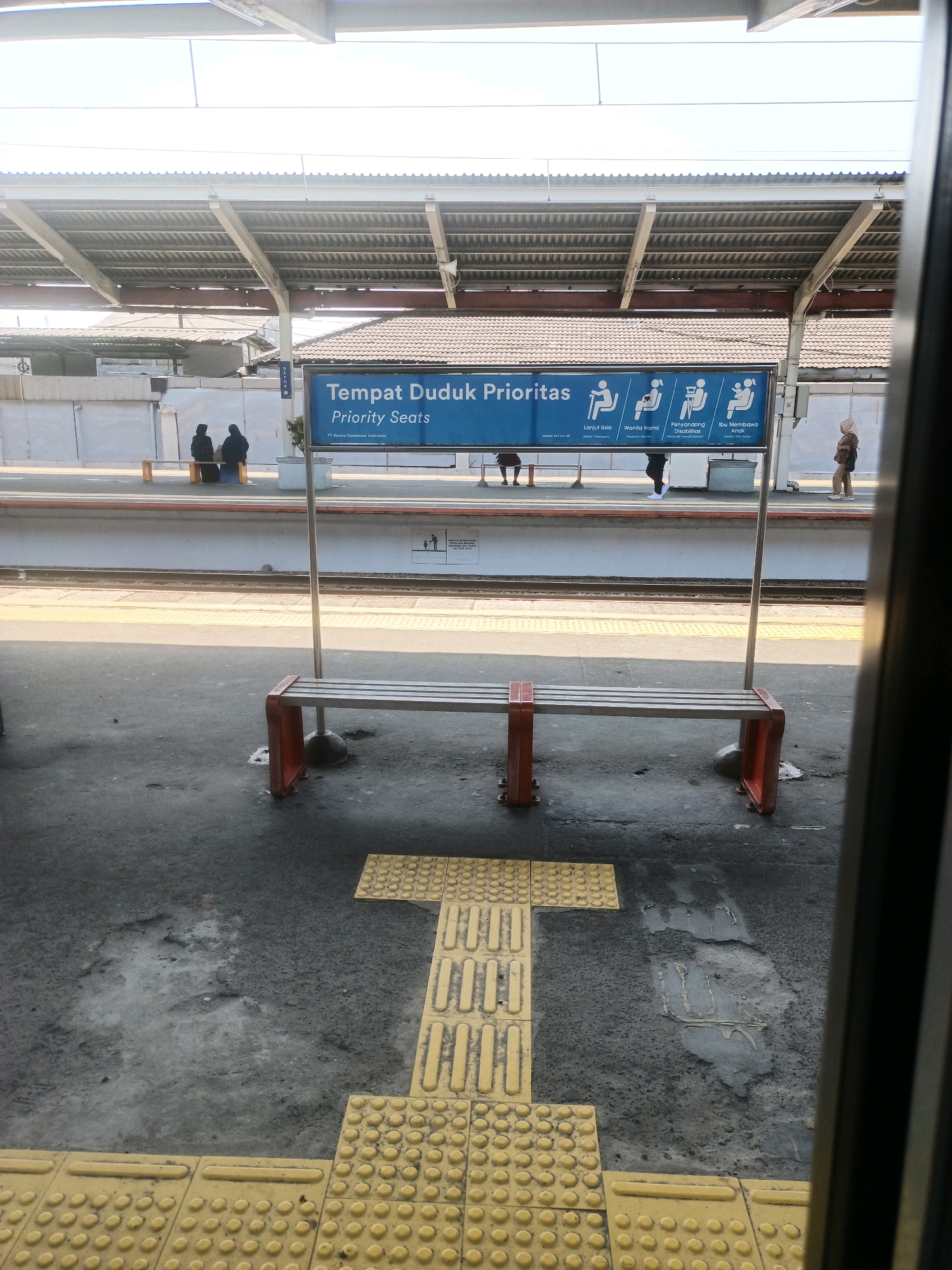 An outdoor photograph taken from inside a train carriage shows a train station platform in Depok, Indonesia. The platform is equipped with tactile paving tiles to assist visually impaired passengers. A blue sign with white text and pictograms indicates "Tempat Duduk Prioritas" or "Priority Seats," designating seating for the elderly, pregnant women, people with disabilities, and those carrying children. Several people are visible on the platform; two individuals are seated on a bench under the priority seating sign, and another person walks by. A metal bench with red supports is positioned in the foreground. The station has a covered roof supported by metal beams and columns.