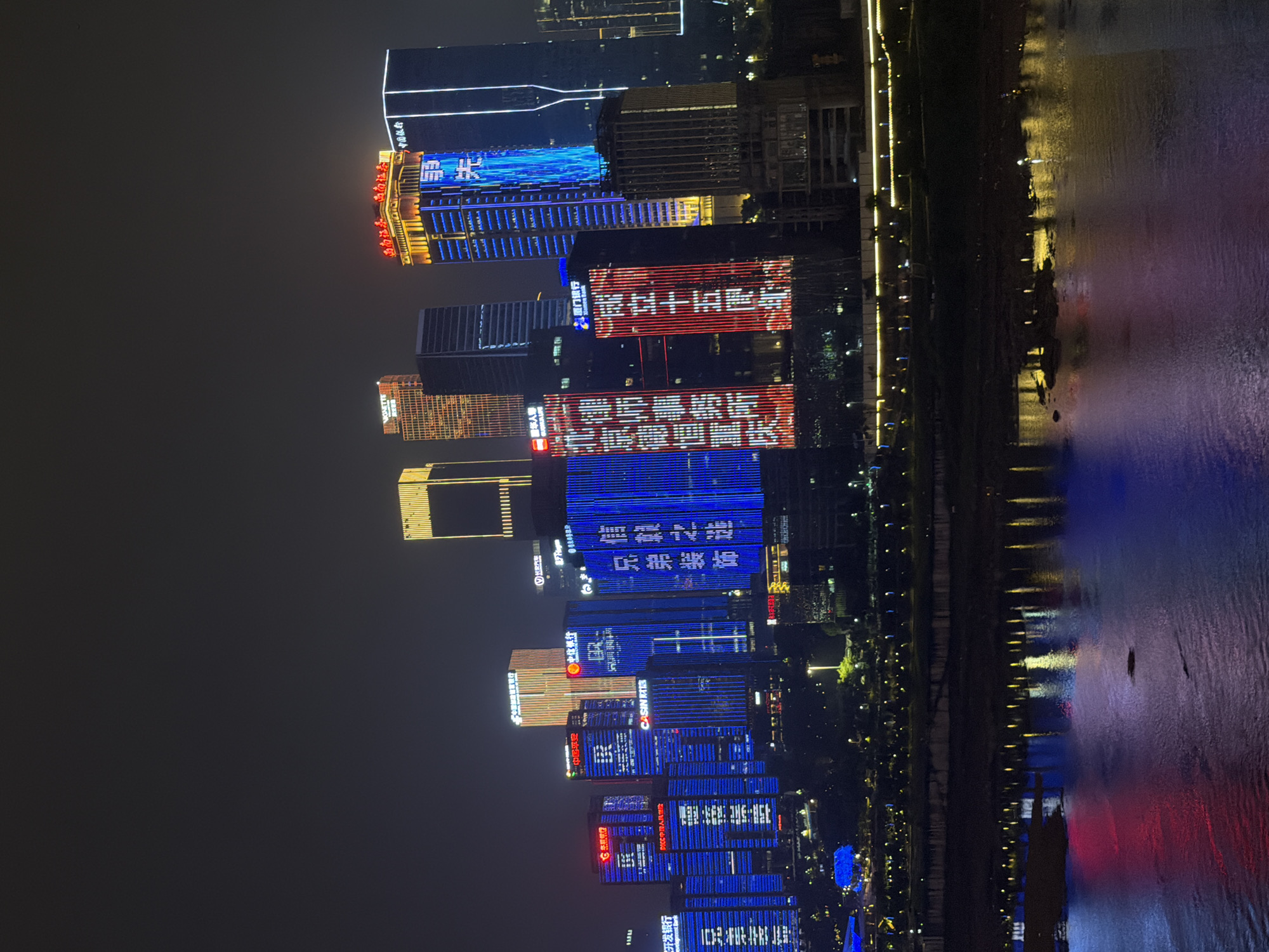 This is a nighttime, exterior, long-shot photograph of a modern city skyline, specifically Yuzhong District, China. The image captures numerous skyscrapers illuminated with vibrant LED lights, displaying advertisements and decorative patterns. The lights range in color, predominantly blue, red, and yellow, creating a dynamic and colorful spectacle against the dark night sky.
The buildings vary in height and architectural design, some featuring distinct vertical or horizontal lighting elements. Several buildings prominently display Chinese characters and logos, suggesting a commercial or financial district. The lower portion of the image shows a body of water, likely a river, reflecting the colorful lights of the city, creating a mirror image on the surface. Along the edge of the water, a well-lit promenade or embankment is visible, lined with streetlights.
There are no discernible people or animals in the frame. The focus is on the architectural grandeur and the illuminated cityscape. The overall atmosphere is one of urban vibrancy and technological advancement.