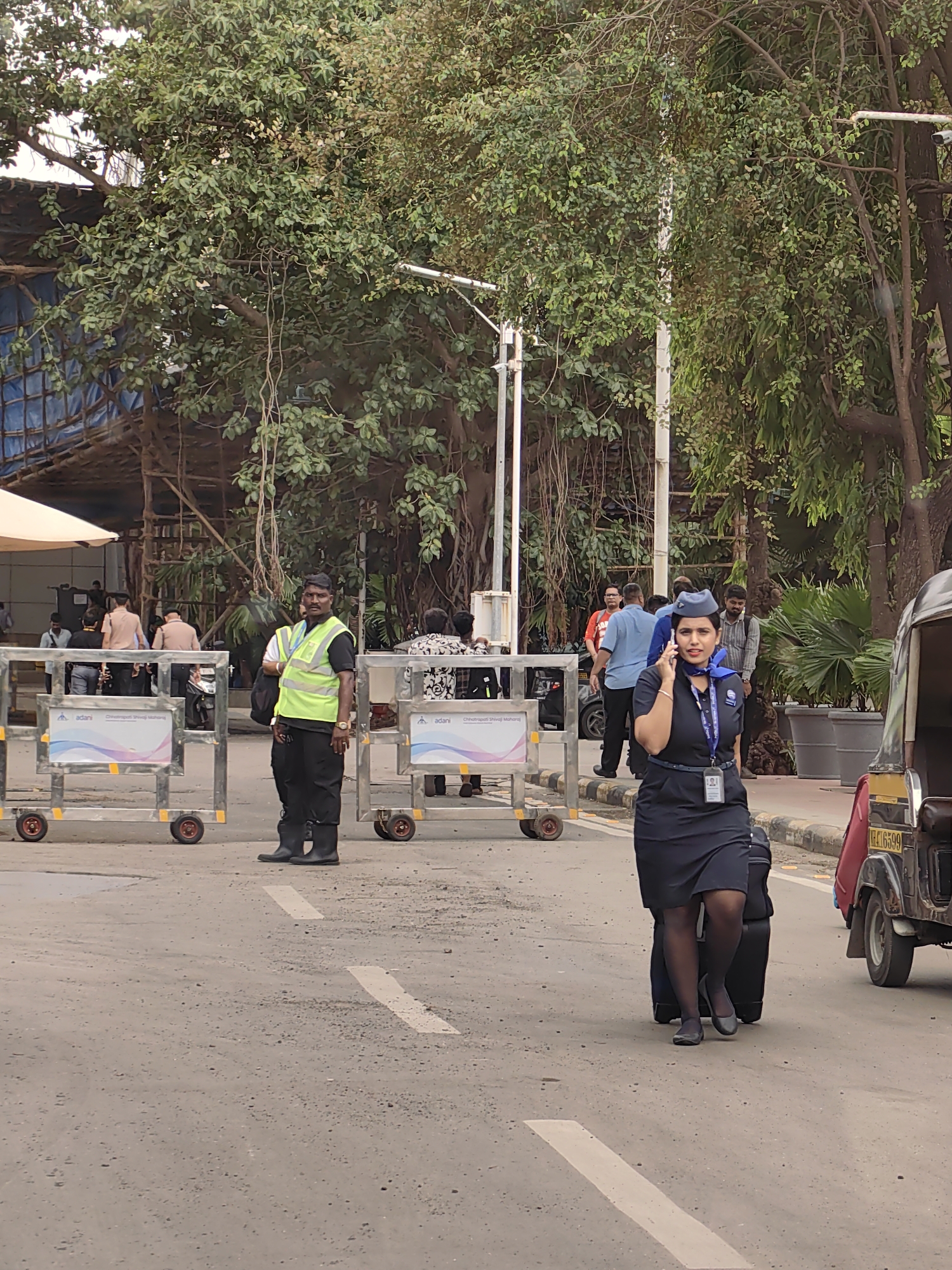 In Mumbai, India, a female flight attendant, wearing a dark blue uniform with a blue scarf, walks on a paved road while speaking on her phone and pulling a rolling suitcase. She is seen from the chest down, with her hair pulled back and a nametag visible on her uniform. Behind her, a male security guard in a bright yellow vest and black pants stands beside metal barriers. Several other individuals are visible in the background, some near a structure with a tarped roof, and trees with dense foliage frame the scene. A three-wheeled auto-rickshaw is parked on the right side of the road. The scene appears to be an outdoor area, possibly near an airport or transportation hub, given the presence of security personnel and the flight attendant.
