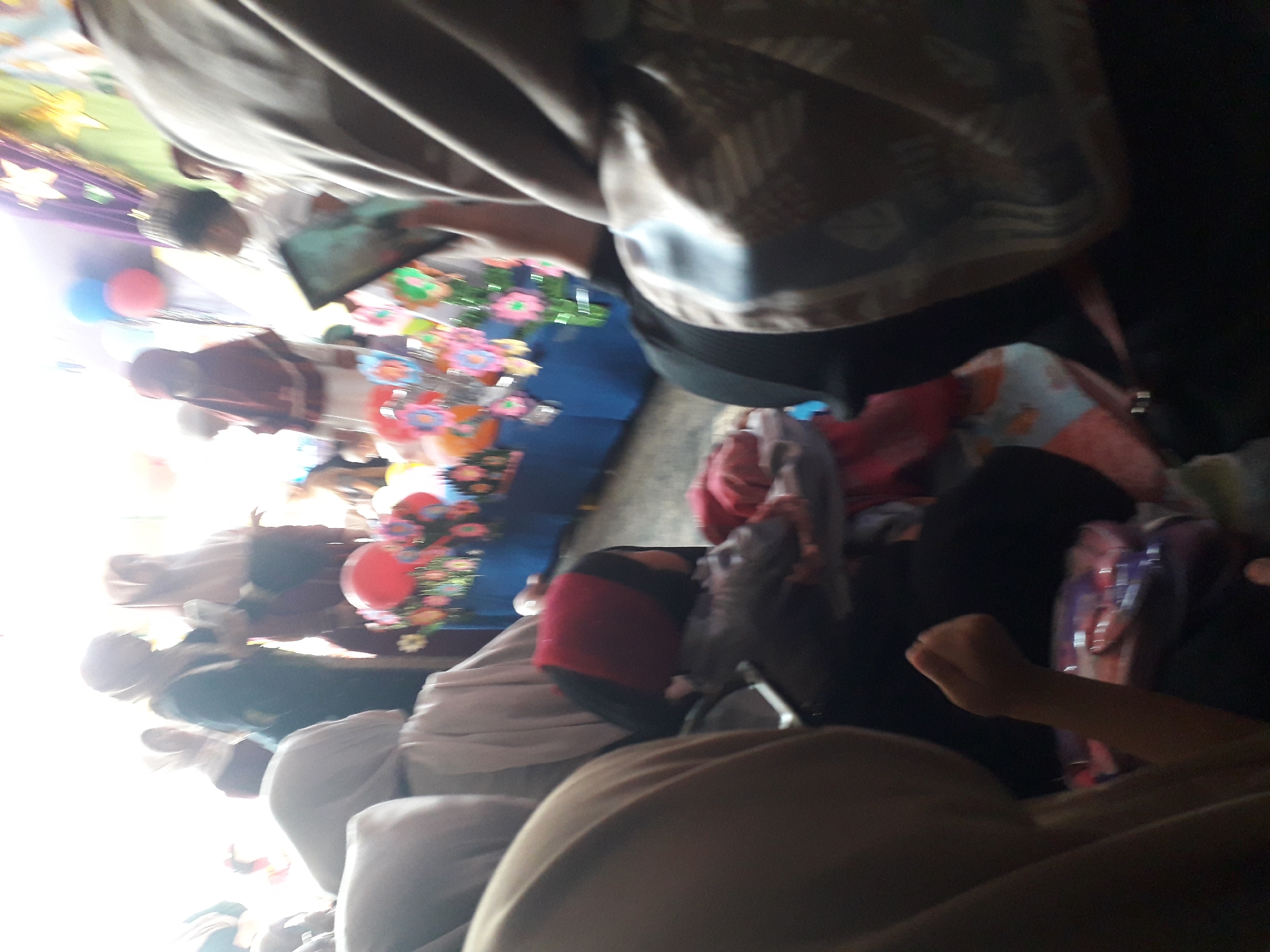 The image appears to be taken indoors at an event, possibly a celebration or a gathering, given the presence of decorations like balloons and colorful paper flowers. Several people are visible, many wearing headscarves, suggesting a cultural or religious context. The lighting is bright, creating a somewhat washed-out effect in some areas, particularly in the background. The foreground is dominated by the backs of several individuals, obscuring much of their faces and specific actions. There's a table or display area with the decorations, and in the mid-ground, a person is holding what looks like a smartphone. The overall impression is of a lively, possibly crowded event. The location in Cankuang, Indonesia, would place this event within a specific cultural and geographical context.