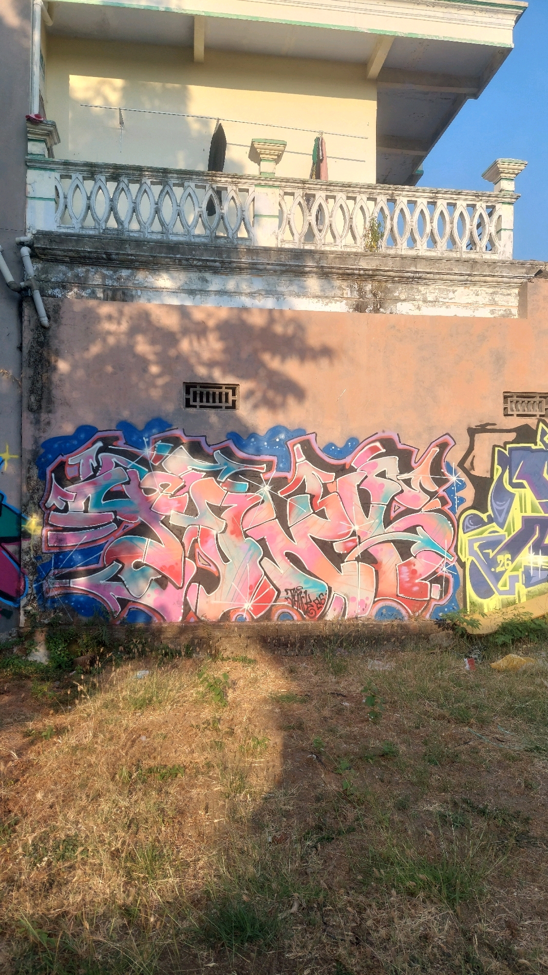 The image shows a section of an outdoor wall in Ronowijayan, Indonesia, covered in graffiti. The wall is painted a pale orange color. A large, elaborate graffiti piece dominates the center of the wall, featuring swirling letters in shades of pink, red, teal, and black, outlined in blue and black. There are white star-like accents scattered throughout this piece. To the right of this central graffiti, another piece is visible, primarily in yellow and purple with some black outlines and a number "26" visible. The wall has a small, dark rectangular vent near the top center. Above the wall is a white, ornate balcony railing with an oval pattern, and a pale yellow building facade. The ground in front of the wall is covered in dry grass and dirt, with some shadows cast from foliage above. The sky is clear and blue.