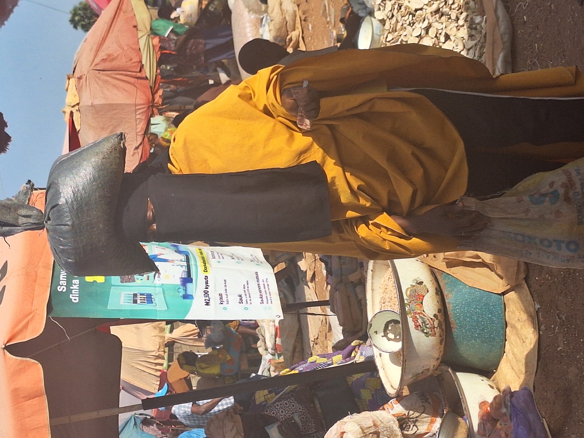 A street scene in Kakuri Gwari, Nigeria. A woman with a black niqab and a mustard yellow robe stands with a black plastic bag on her head, holding what appears to be currency in her hand. Beside her, there are several enamel basins filled with various goods, possibly for sale. Above her, there's a banner advertising services and prices in local currency. In the background, there is a variety of materials, and a mix of other people. The scene is characterized by the busy, crowded atmosphere of a marketplace.