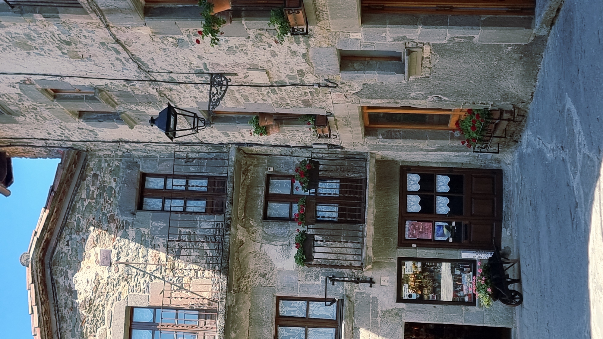This is a medium shot, exterior, eye-level photograph taken in Rupit, Spain, showcasing a narrow cobblestone street lined with ancient stone buildings. The sun is bright, casting shadows and illuminating the weathered stone facades. On the left, a multi-story stone building features a wrought-iron balcony on its upper level, adorned with potted red flowers. Below, two dark wooden doors are visible, one with frosted glass panes and the other with multiple small, white lace curtains. To the left of the doors, a shop window displays various items. In front of the shop, a black, vintage wheelbarrow filled with pink flowers is parked on the cobblestones.
The building on the right is also constructed of rough-hewn stone. It has a large, dark wooden door with a decorative metal handle and a window above it. A black wrought-iron lantern hangs from the wall to the right of the door, its design ornate. Flower boxes filled with red blooms are strategically placed on ledges and balconies on this building as well. The cobblestone street appears wet in places, with a light reflecting off the surface. The overall impression is one of historic charm and rustic beauty.