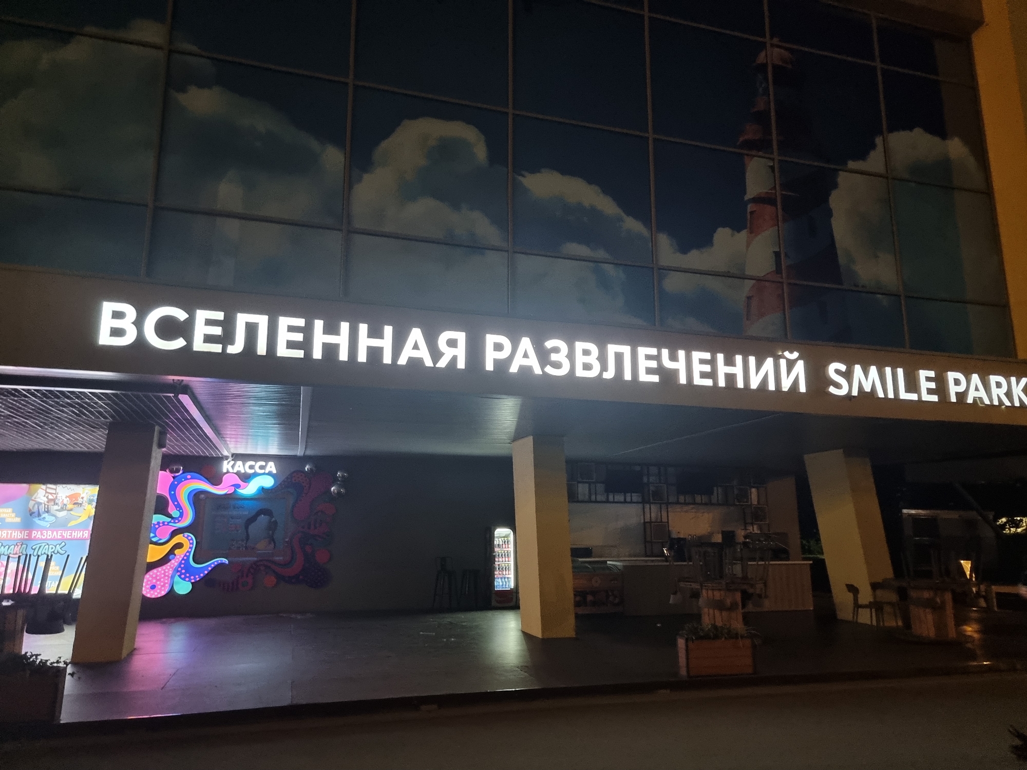 The image shows the exterior of a building at night, likely an entertainment complex. A large, illuminated sign across the facade reads "ВСЕЛЕННАЯ РАЗВЛЕЧЕНИЙ SMILE PARK" (Universe of Entertainment SMILE PARK). The building's facade is predominantly glass, with a large mural depicting clouds and a lighthouse visible through it.
Below the main sign, there is a covered entrance area. To the left, a colorful mural with abstract designs and the word "КАССА" (Cashier) is visible. There are also advertisements for attractions with Russian text that includes "УПАТНЫЕ РАЗВЛЕЧЕНИЯ" and "МАЙЛ ПАРК". A vending machine for drinks is situated further inside the covered area. Tables and chairs are visible, some stacked on top of others, suggesting the establishment may be closed or preparing for closure. The ground is wet, indicating recent rain. The overall impression is of a leisure facility located in a city. While the city is identified as Moscow, Russia, there are no specific landmarks or contextual clues within the image to pinpoint an exact location within Moscow.