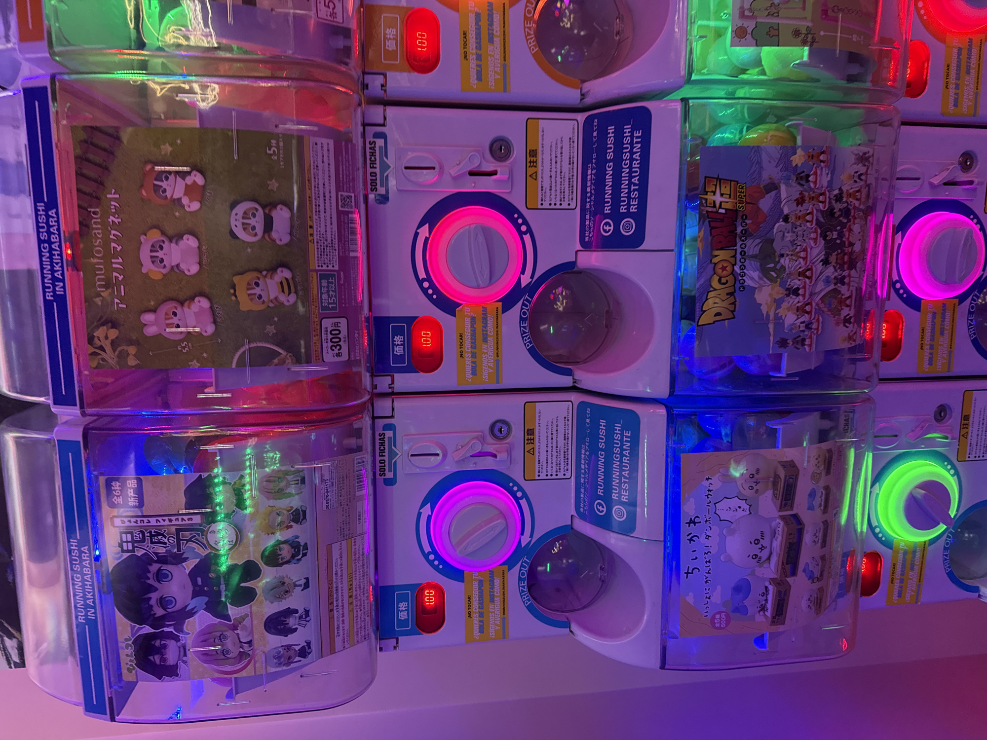 The image depicts a close-up of several gashapon (capsule toy) machines, likely located in Japan, judging by the Japanese text and the types of toys. The machines are stacked and feature various anime and manga-themed merchandise, including "mufosand" animal magnets, "Dragon Ball Super" figurines, and "Chiikawa" items. Each machine has a price display (100 or 300 yen), a coin slot, and a capsule dispensing area. The lighting is vibrant and colorful, with neon rings around the dispensing areas in colors like pink, green, and blue. Promotional signage around the machines includes warnings and instructions in Japanese, as well as social media callouts for "Running Sushi." There is no indication that this is anywhere in Madrid, Spain.