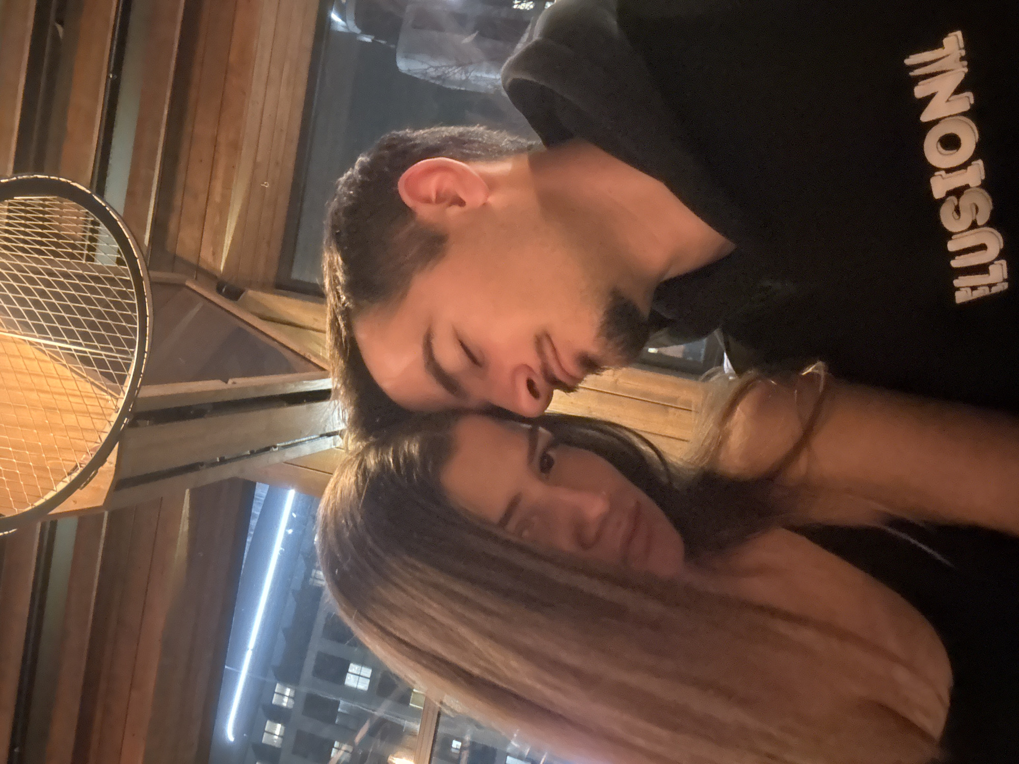 A close-up shot depicts a man and a woman embracing indoors, likely in a bar or restaurant due to the wooden interior. The man is wearing a black hoodie with the word "ILLUSION!" printed vertically on it. He has short dark hair and a trimmed beard. His eyes are closed. The woman has long blonde/brown hair. Her eyes are open and directed towards the camera. Outside the window, city lights indicate an urban setting, possibly in Sofia, Bulgaria. A metallic lighting fixture is visible in the upper left corner of the frame. The lighting suggests an intimate and warm environment.