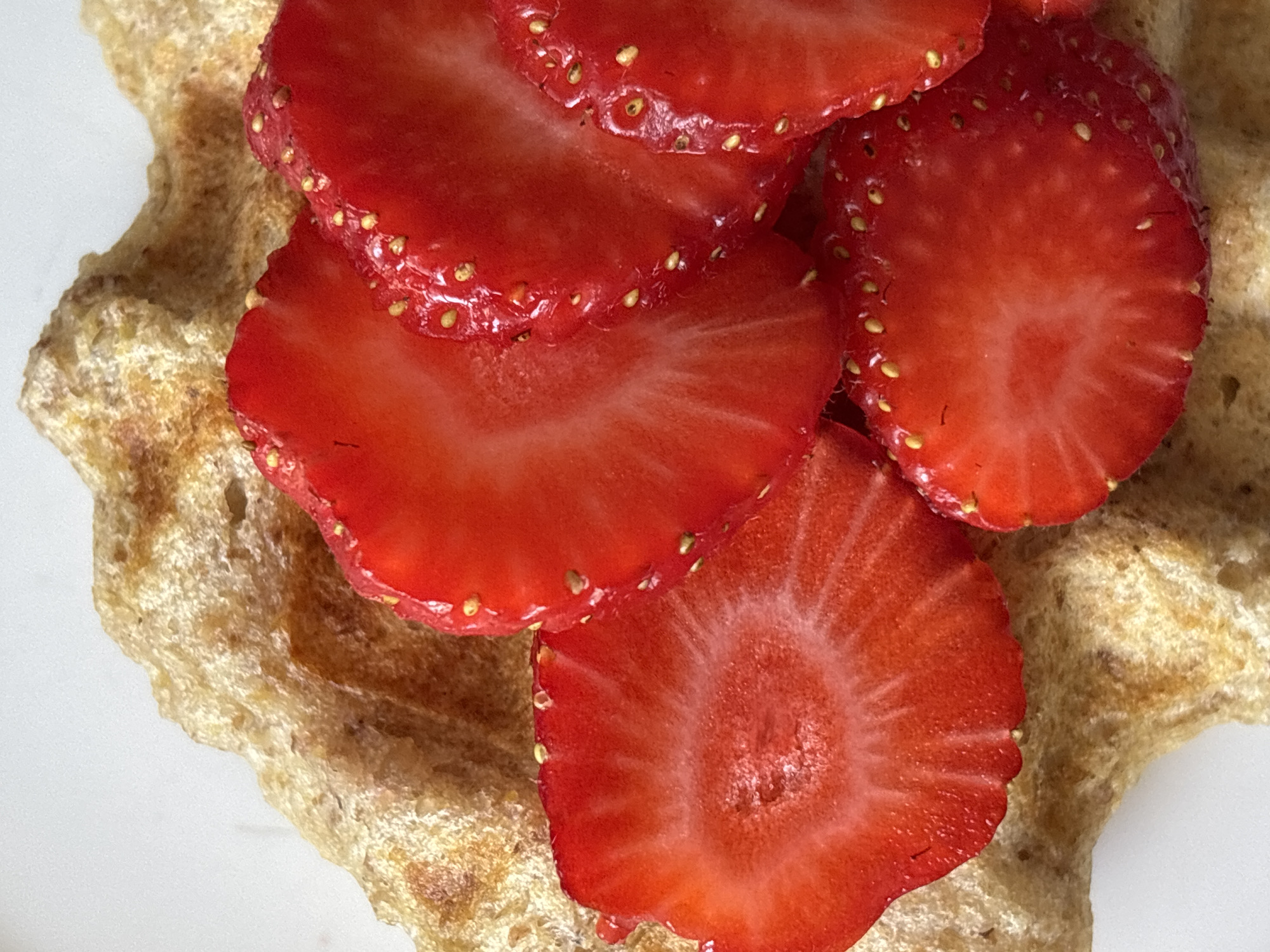 The image is a close-up, overhead shot of sliced strawberries on a piece of what appears to be a waffle or pancake. The strawberries are bright red with visible seeds and a textured surface. The waffle or pancake has a light brown, slightly porous texture. The background is plain white. The image is a food photograph and does not depict any location in Sochi, Russia.