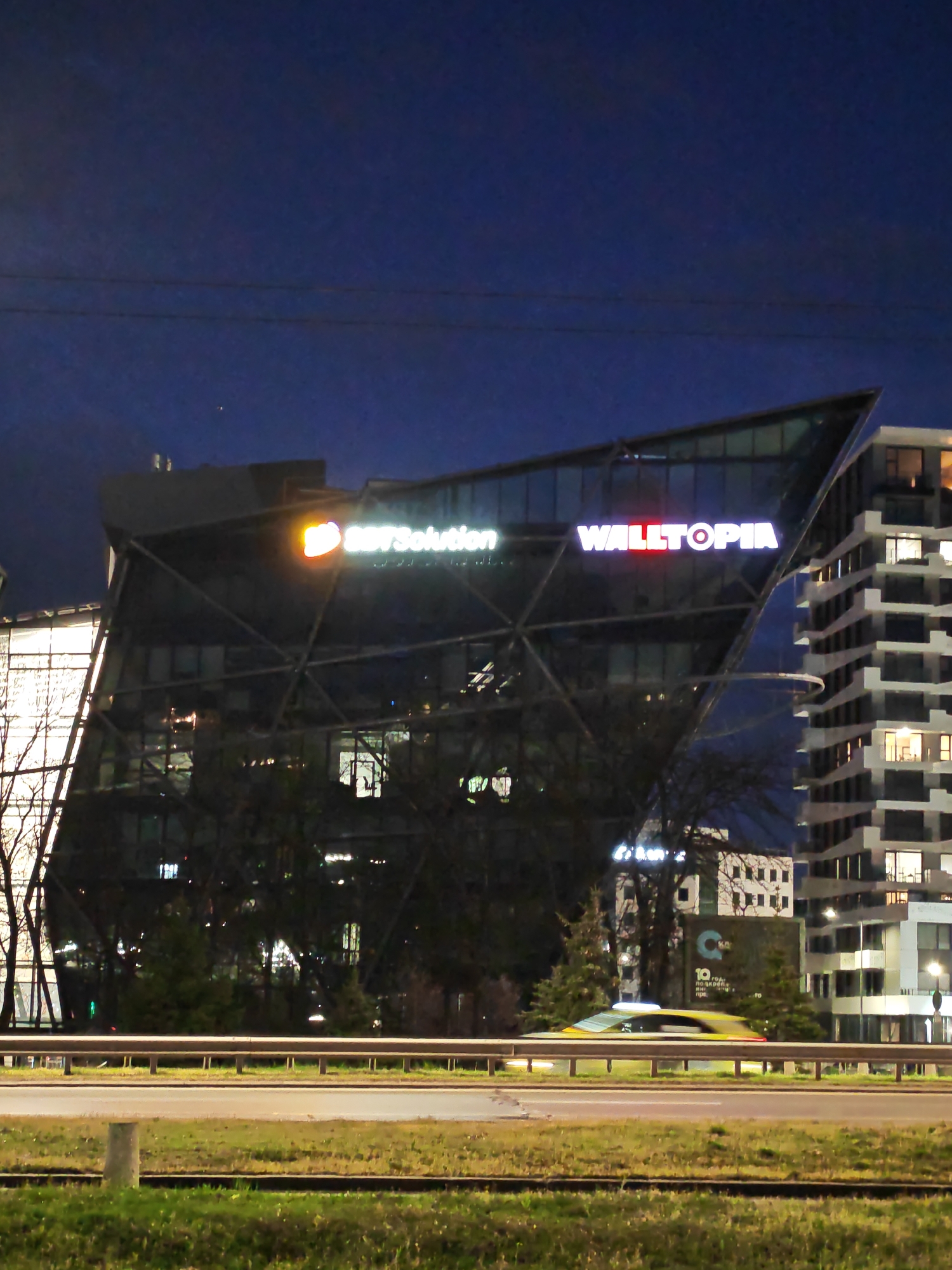At night, an angular glass and steel building stands, bearing the illuminated signs of "DP Solution" and "WALLTOPIA." Adjacent to it, a taller, modern building features multiple lit windows. A multi-lane road separates the buildings from a grassy area. A yellow car drives past on the road. The image is taken in the city of Sofia, Bulgaria, as evidenced by the signage on the building.