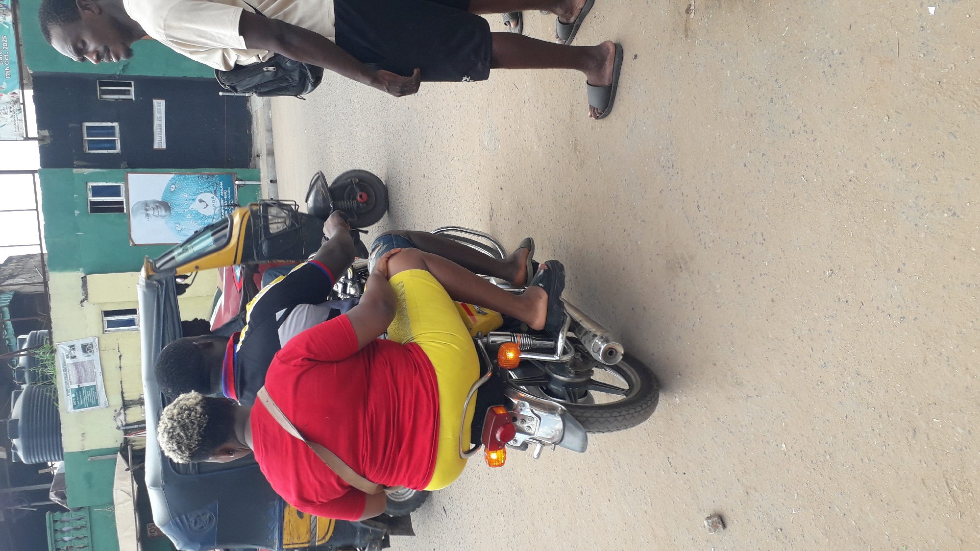 The image depicts a scene in Onitsha, Nigeria. Three individuals are on or near a motorcycle-drawn auto rickshaw. The person in the foreground wears a red shirt and yellow shorts, sitting on the back of the motorcycle. The person in front of them wears a black and blue top and shorts. The rickshaw driver is visible in front of the passengers. A fourth person is visible in the upper part of the image, walking and wearing a light-colored T-shirt and black shorts. Buildings are visible in the background.