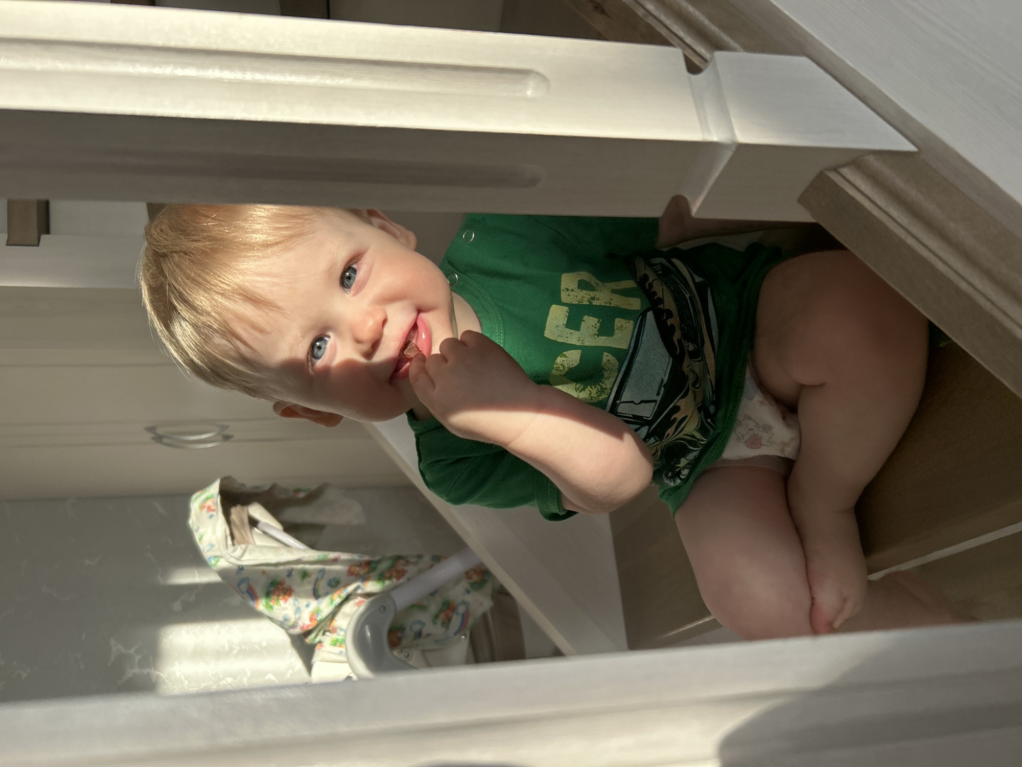 A toddler with blond hair and blue eyes wearing a green shirt and a diaper sits under a structure of white wooden beams. The child is holding something small near their mouth, possibly food. A baby carrier is visible in the background. The photograph was taken indoors. There is no specific information in the image to confirm its location in Chaikovsky, Russia.