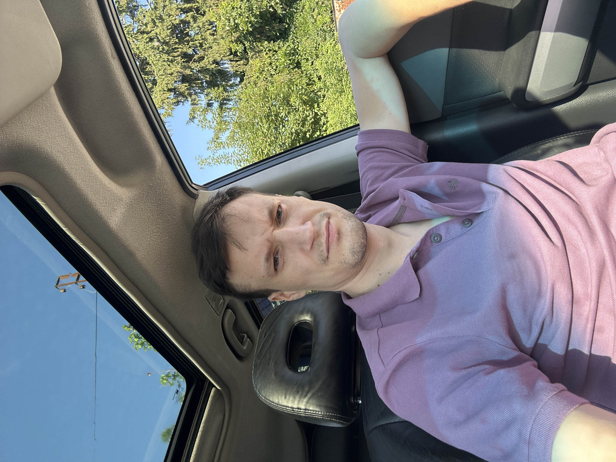 In Shatava, Ukraine, a man sits in the driver's seat of a car, looking towards the camera with a slight grimace. He is wearing a purple polo shirt. The car's sunroof is open, revealing a bright blue sky and green trees outside. The interior of the car is visible, including the black leather seat and headrest. The man's right arm rests on the car door, and sunlight casts a shadow on his shirt. The scene appears to be captured on a clear, sunny day.