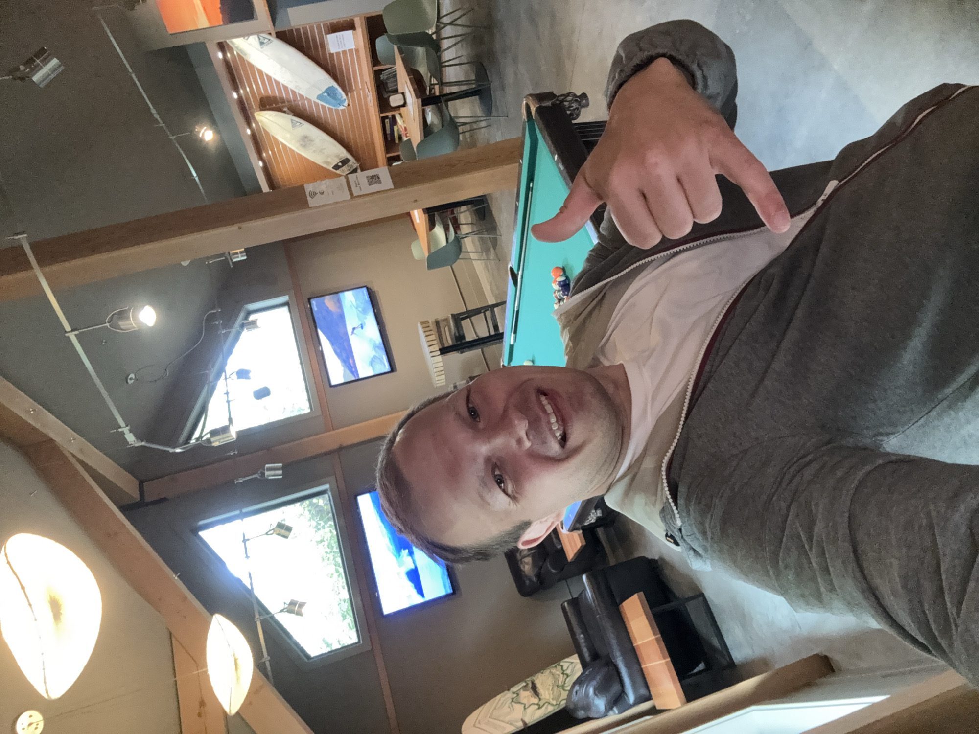 The image is a tilted selfie of a man in a gray jacket and white shirt, making a "shaka" hand gesture towards the camera, appearing to be taken inside a recreational establishment in Tofino, Canada. In the background, a pool table with billiard balls is partially visible, along with surfboards mounted on the wall, a dining area with tables and chairs, and multiple television screens displaying images of the ocean. The interior features a wooden beam structure with modern lighting.