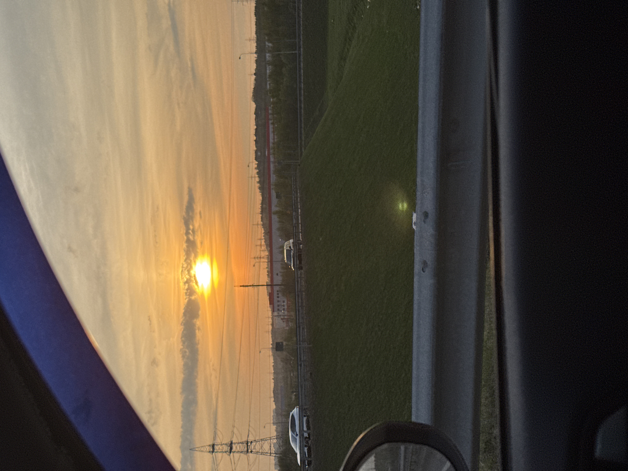 The image depicts a sunset viewed from inside a vehicle, possibly on a highway near Poroshkino, Russia. The sun is setting towards the left side of the frame, casting a warm, golden light on the sky. In the foreground, there is a glimpse of the vehicle's interior, including the side mirror and part of the dashboard. Outside the vehicle, there is a grassy area bordering the road. Power lines and towers are also visible, silhouetted against the setting sun. Further in the distance, faint outlines of buildings and infrastructure suggest a suburban or peri-urban environment. Cars are visible on the road.