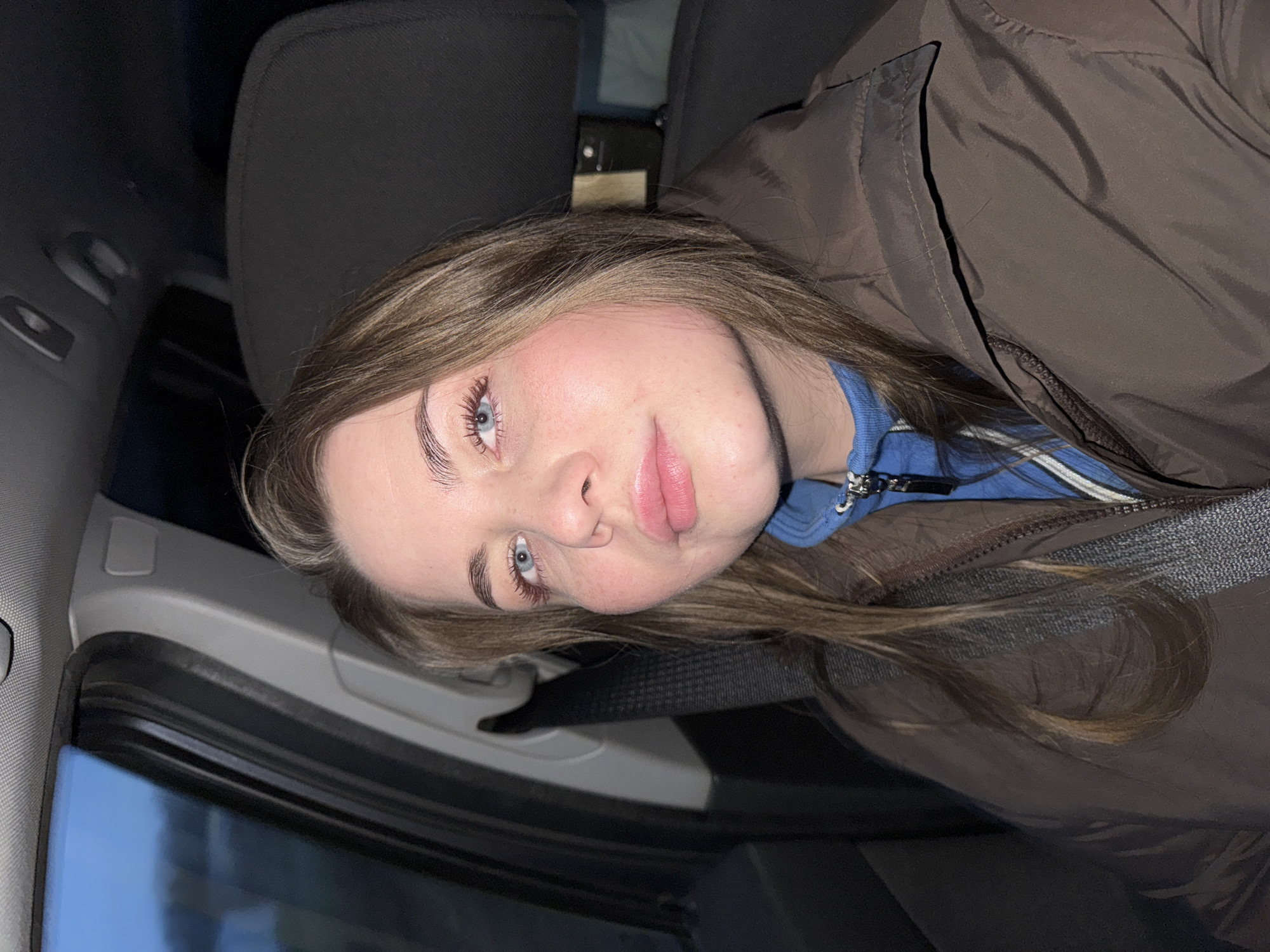 A young woman is depicted in a close-up portrait, seated inside a car in Ol'khovochka, Russia. She has long, light brown hair that cascades over her shoulders and striking blue eyes that look directly at the viewer. Her complexion is fair, and her lips are a soft pink. She is wearing a dark brown, possibly padded, jacket over a blue top, which features some white stripes or detailing around the zipper area. A seatbelt crosses diagonally over her chest.
The setting is the interior of a vehicle, likely a car, with dark gray and black fabric and plastic components visible in the background, including a headrest and parts of the door frame and dashboard. The scene outside the car window is dark, indicating that the photo was taken at night or in the late evening. The woman's expression is calm and neutral, with a slight tilt of her head. There is no visible text in the image.