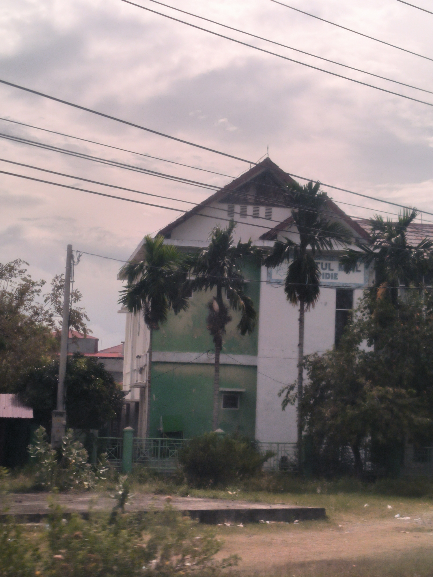This daytime image captures an outdoor scene in Lampeudeu Baroh, Indonesia, featuring a multi-story building, likely an institutional or school facility, under an overcast sky.
The central subject is a white building with a prominent, dark-tiled pitched roof. The lower part of its facade is painted a distinct green, contrasting with the white upper section. Several tall palm trees are positioned in front of and around the building, partially obscuring its details. Other smaller trees and bushes are visible in the mid-ground and background.
In the immediate foreground, there is an unkempt area of sparse vegetation and dirt, traversed by a low concrete curb or platform. A green metal fence acts as a boundary in front of the building. To the left, a utility pole rises, from which multiple black power lines stretch horizontally across the upper part of the frame.
The sky is cloudy and grey, indicating an overcast day with soft, diffused lighting and no harsh shadows. No specific activity or people are visible in the static scene.
Visible text on the building's facade, positioned between two palm trees, appears in blue lettering. Fragments of the text are legible, reading "FAKULTAS" (partially visible on the left), "UL ML" on the upper line, and "PIDIE" on the line below.