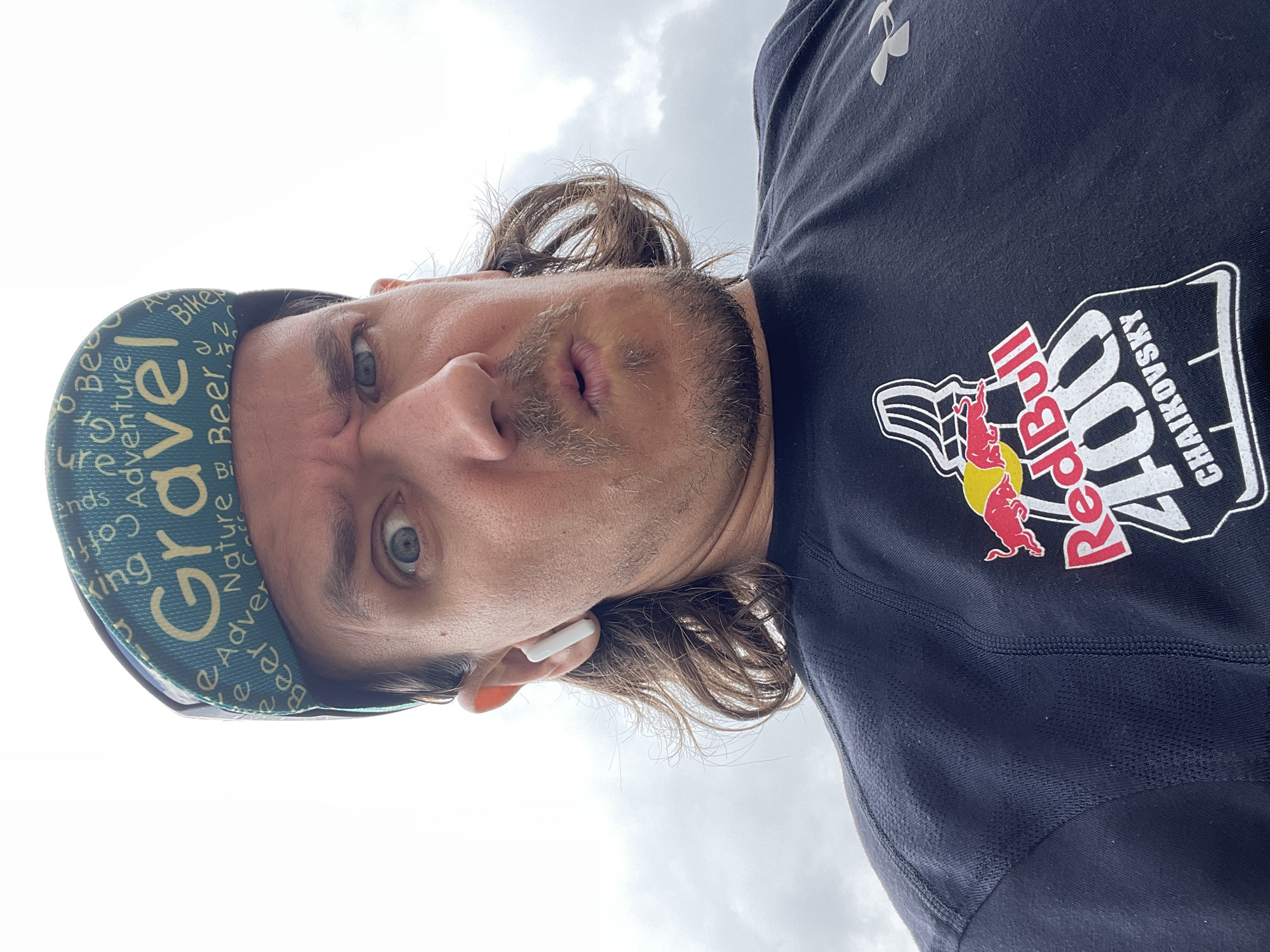 An extreme close-up, low-angle selfie shows a male subject with light brown, medium-length hair, light stubble, and blue eyes. He wears a surprised or exaggerated expression with slightly raised eyebrows, widened eyes, and parted lips. One white earbud is visible in his right ear.
He is wearing a blue/teal cycling cap patterned with words such as "Gravel," "Adventure," "Beer," "Nature," "Bike," "Friends," and "Coffee." His dark blue or black t-shirt features a prominent "Red Bull" logo with a charging bull, and text that reads "400 CHAIKOVSKY." An Under Armour logo is also visible on the upper left chest area of the shirt.
The background is an overcast or cloudy sky, indicating an outdoor setting during daytime. The perspective suggests the photo was taken from below the subject, possibly during or after an outdoor activity related to cycling or an endurance event, given the apparel and branding.