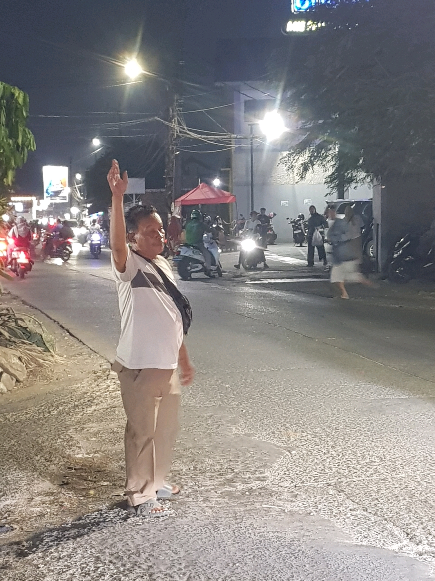 In Setiamekar, Indonesia, at night, a man stands on a street. He is wearing a white t-shirt with a dark stripe across the chest, light brown pants, and grey slide sandals. His right arm is raised, with his hand open. He has a dark bag slung across his body. The street is paved with asphalt and has some debris along the side. Several motorcycles and a few people are visible in the background, some on motorcycles, others walking. There are also lights from buildings and streetlights contributing to the nighttime illumination. A red tent structure is also visible in the background.