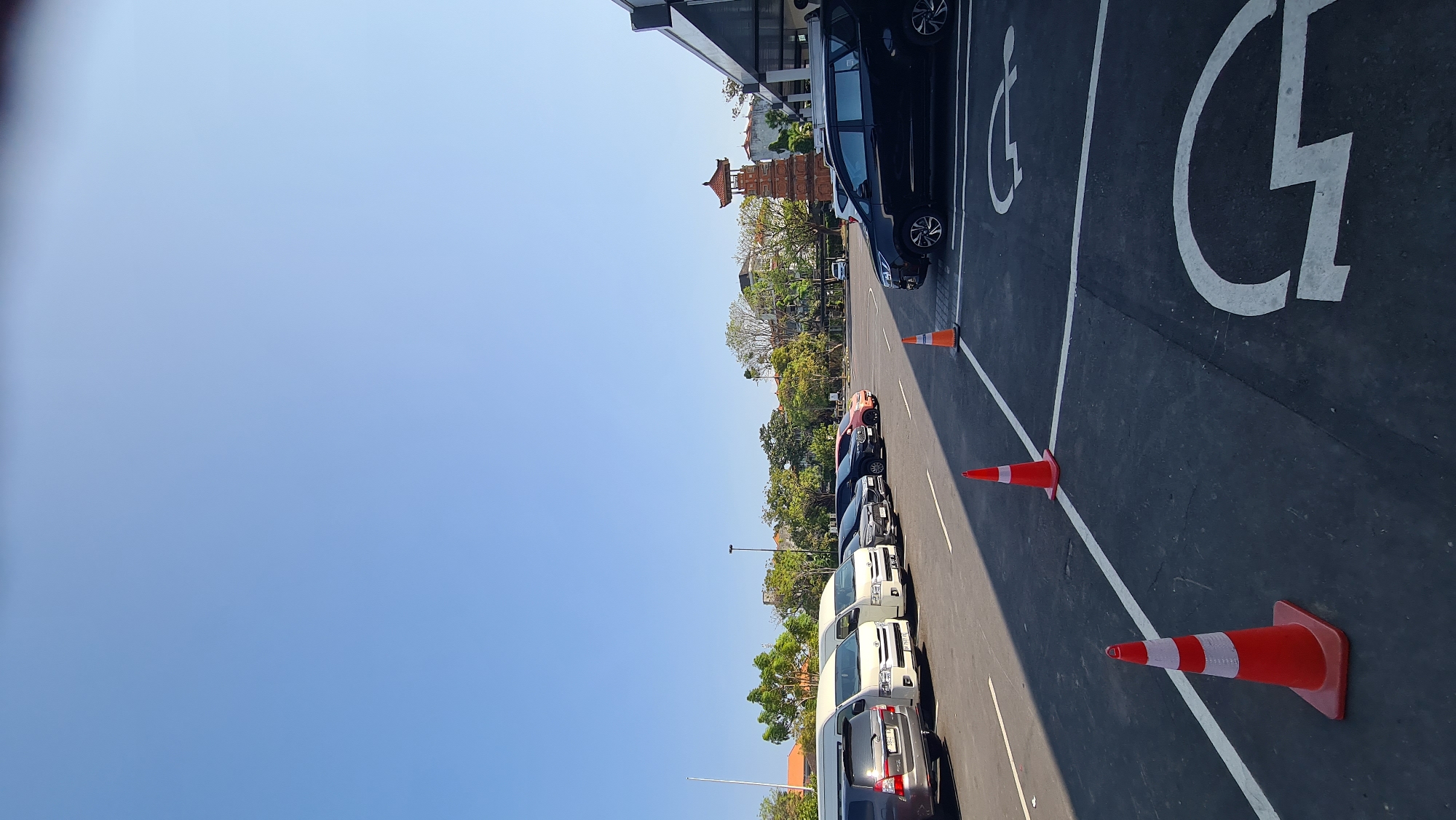 A clear, cloudless blue sky dominates the upper half of the frame. The lower half depicts an outdoor parking lot with an asphalt surface, marked with white lines. Several vehicles are parked in the lot, including vans and SUVs of various colors. Orange traffic cones are strategically placed throughout the parking area, with two in the foreground and one further back. In the mid-ground, a distinctive multi-tiered, pagoda-style tower with a red roof stands against a backdrop of green trees and buildings. The architecture suggests an Indonesian setting, likely in Denpasar, Indonesia, given the cultural motifs of the tower. The scene appears to be a bright, sunny day, possibly in the morning or afternoon, indicated by the long shadows cast by the cones and cars. There are no people visible in the image. The parking lot has designated spaces, including what appear to be disabled parking spots marked with white wheelchair symbols. The overall scene is one of an organized, functional public space.
