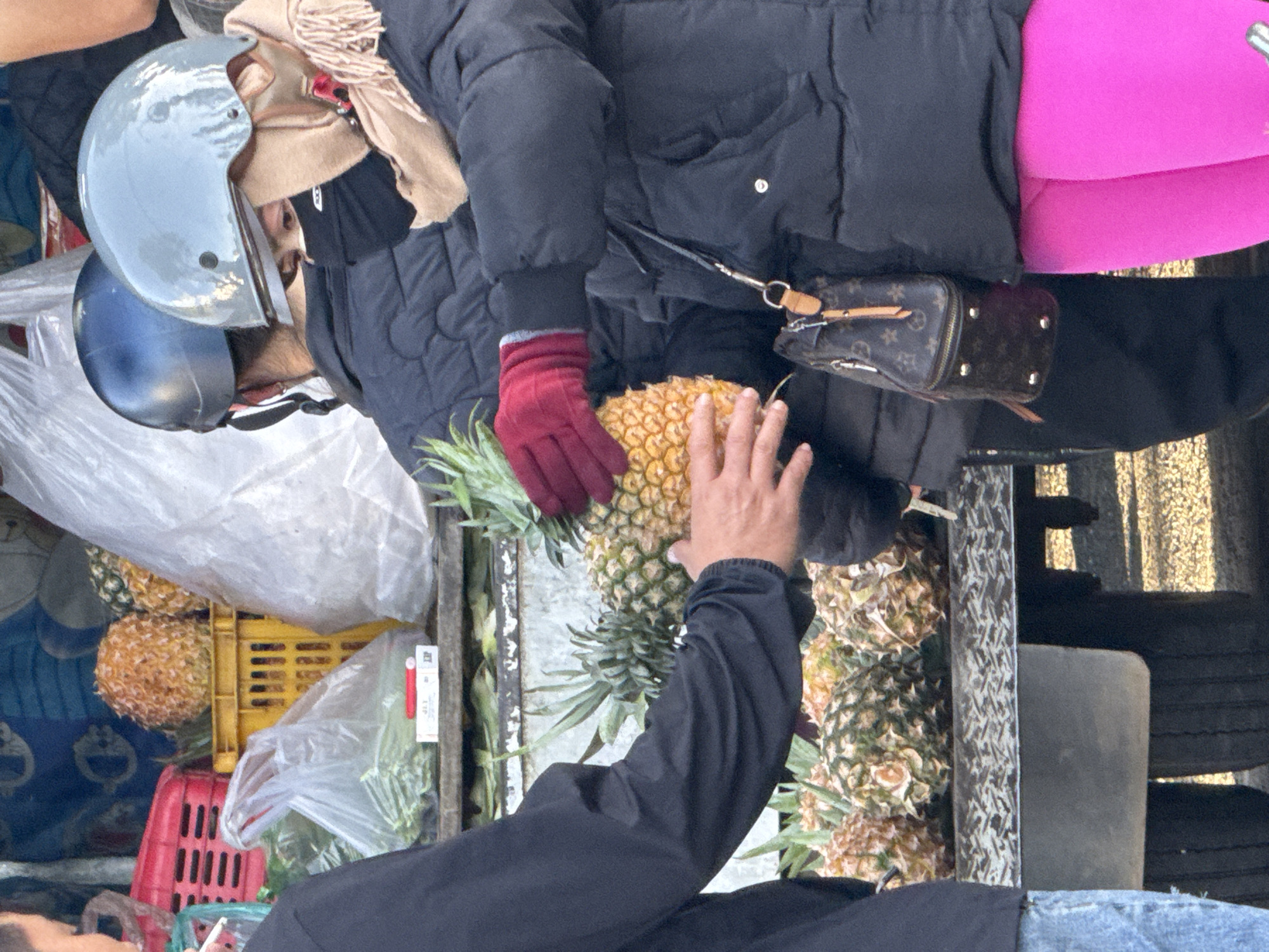A street vendor is selling pineapples from the back of a truck in Da Lat, Vietnam. Two individuals, likely customers, are interacting with the vendor and the fruit. The person on the left, partially visible, is wearing a black jacket and blue jeans and appears to be smoking. The person in the center is wearing a blue helmet and a black puffer jacket, holding a pineapple with a gloved hand. The person on the right, wearing a light blue helmet, a black face mask, a beige scarf, and a black puffer jacket, is also reaching for a pineapple. The truck bed is filled with pineapples and other produce, including what appears to be green vegetables in plastic bags. In the background, there are red and yellow plastic crates and a large white plastic bag. The scene is lit by natural daylight.
