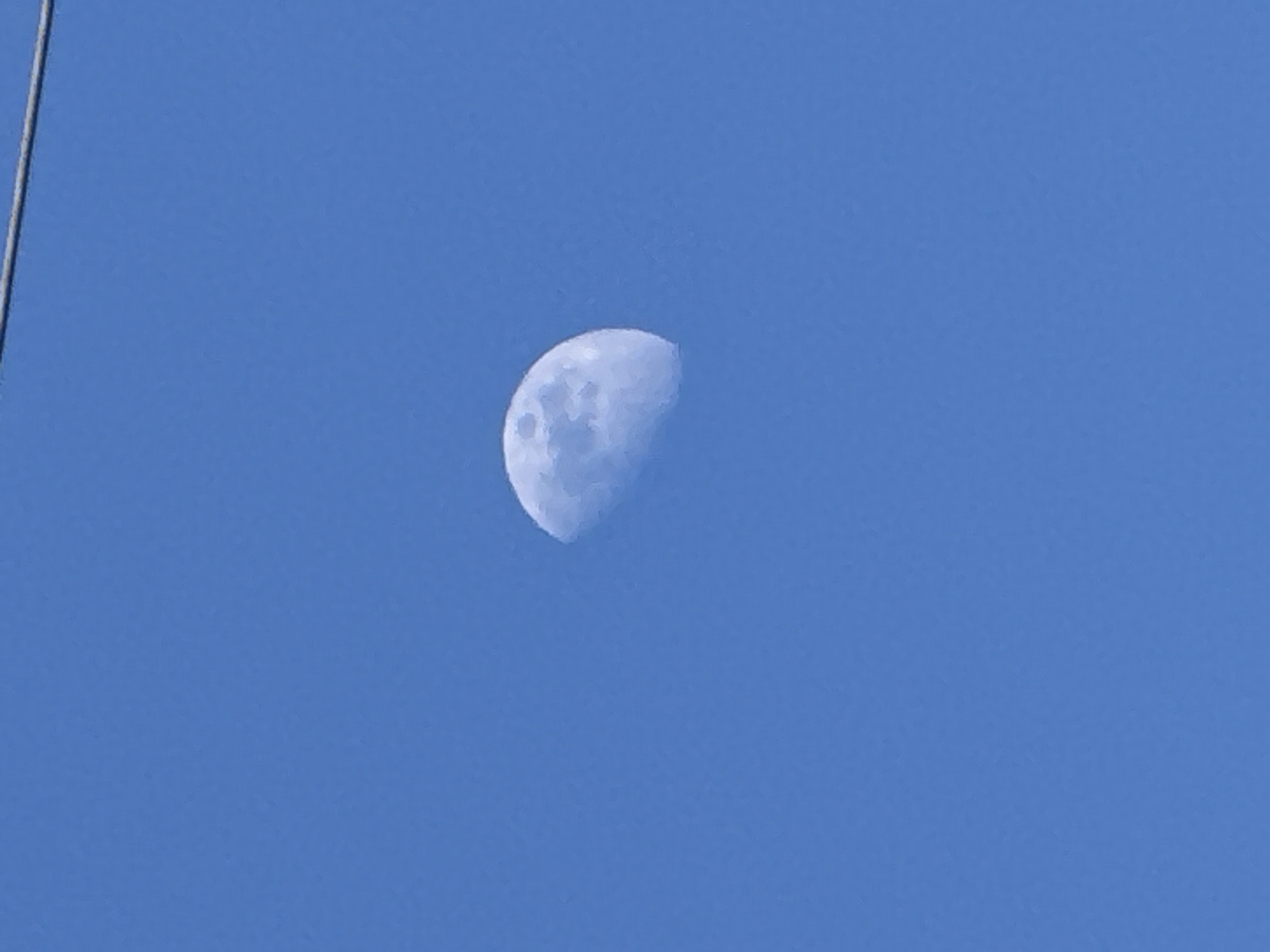 The image displays a clear blue sky during daylight hours, with the Moon visible. The Moon is in a half-moon phase, showing some surface details and craters. The sky is a vibrant, uniform blue, indicating clear weather without clouds. In the upper left quadrant, a thin, light-colored diagonal line, possibly a wire, cable, or antenna, is partially visible against the sky. There are no people, other objects, or text present in the frame. The scene is a serene view of the sky, likely taken from Villebon-sur-Yvette, France, during the day.