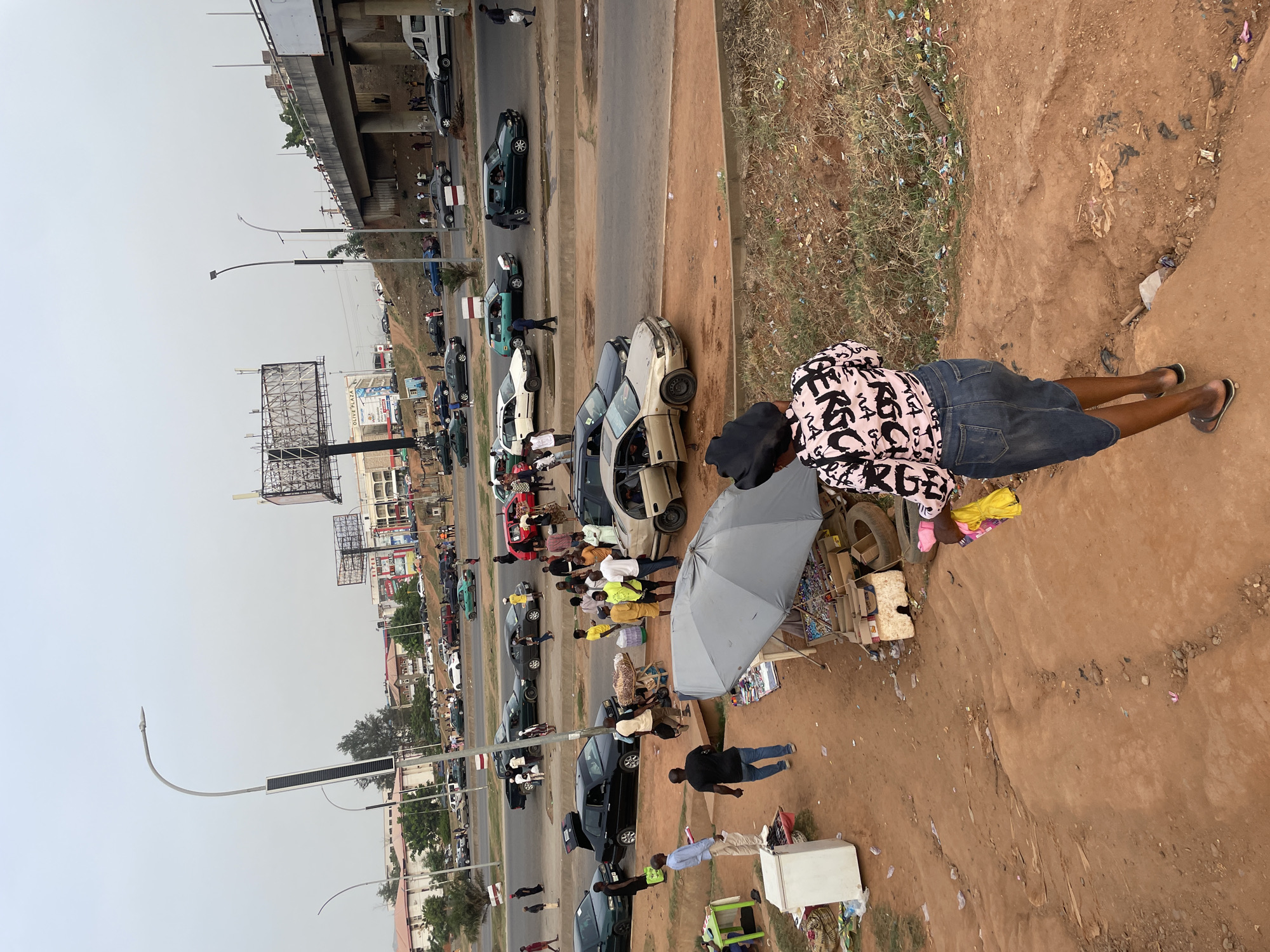 This image captures a busy, urban street scene in Abuja, Nigeria, seen from an elevated perspective. The sky is overcast or hazy, indicating daytime.
In the foreground, a person wearing a pink and black patterned shirt and denim shorts stands on a reddish-brown dirt path, facing away from the camera. This path, littered with debris and grass patches, slopes down towards a main road. Numerous people are visible along the roadside and on the dirt path.
The main road is bustling with traffic, featuring various cars, including sedans, SUVs, and vans, some appearing to be parked or involved in informal commercial activities as doors are open and people are gathered around them. Vendors operate along the side of the road, with one prominent grey umbrella providing shade over a makeshift stall displaying goods. Other informal market setups include pushcarts and items displayed directly on the ground, such as a white refrigerator-like object and discarded tires.
In the midground and background, the road continues, flanked by buildings of varying sizes and styles. Several large billboards are visible, some with structural elements, and one sign in the distance reads "LOUSE PLAZA." Modern streetlights, some equipped with solar panels, line the road. An elevated road or overpass stretches across the upper left portion of the image. The overall scene depicts a vibrant street environment characterized by commerce, pedestrian movement, and vehicle traffic. The pattern on the shirt of the person in the foreground contains abstract, repeated letter-like shapes, such as "MA" or "NA."