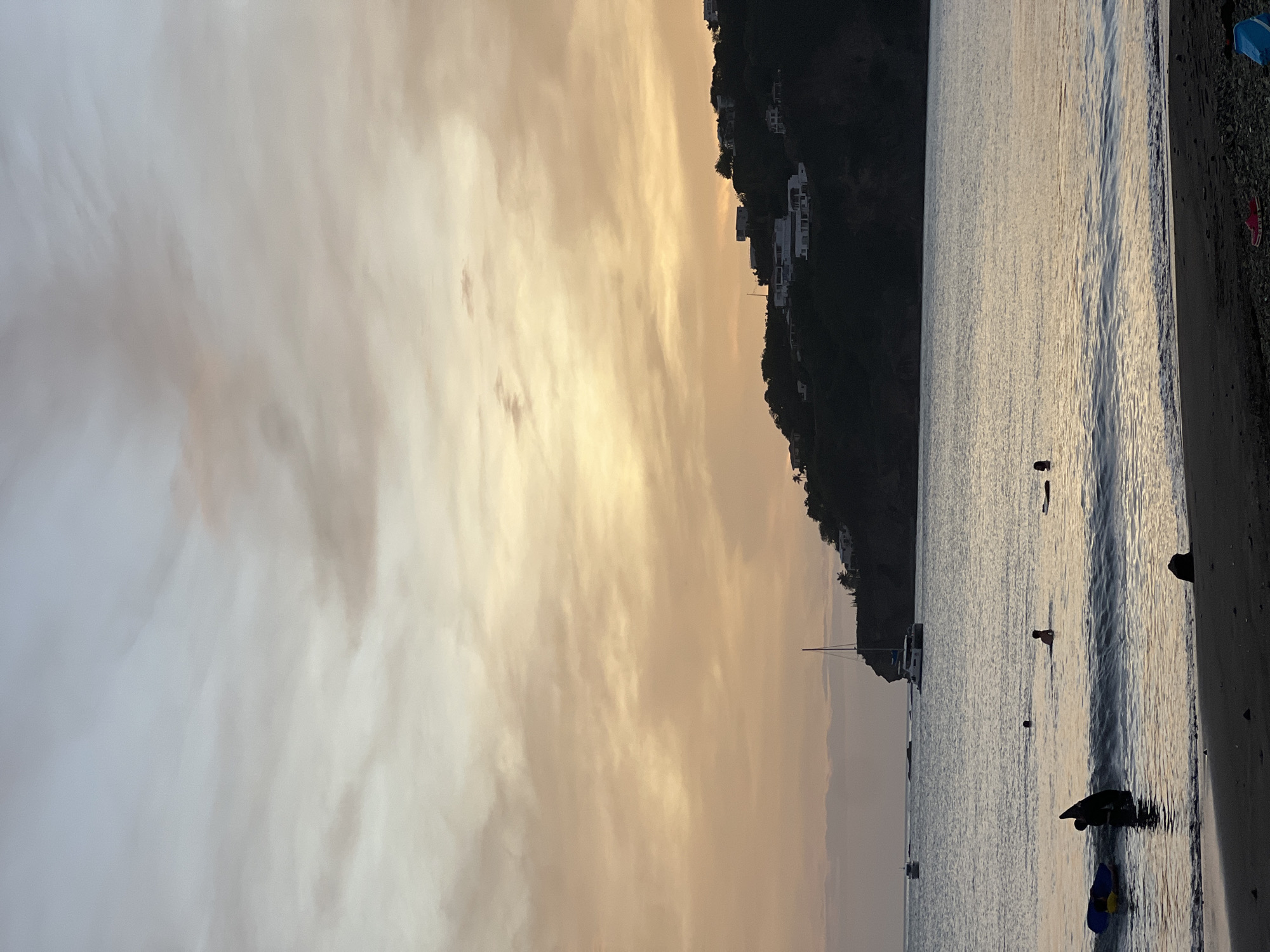 The image captures a serene evening at a beach in San Juan del Sur, Nicaragua. The sky is painted with soft hues of orange and yellow, transitioning into a pale blue, indicating either sunrise or sunset. Wispy clouds are scattered across the horizon. The calm ocean water reflects the sky's colors, with gentle waves lapping at the dark sand beach.
In the foreground, a few individuals are visible in the water. Two people are near the shore, one appearing to be standing and interacting with a blue floatation device, while another person is also in shallow water. Further out, several other figures are present, some swimming and one lying on a floatation device.
A sailboat with a white hull and a tall mast is anchored in the bay, its sail down. Another smaller boat is visible further in the distance. The background features a lush, dark green hillside dotted with white buildings, suggesting houses or villas. The overall atmosphere is peaceful and tranquil.