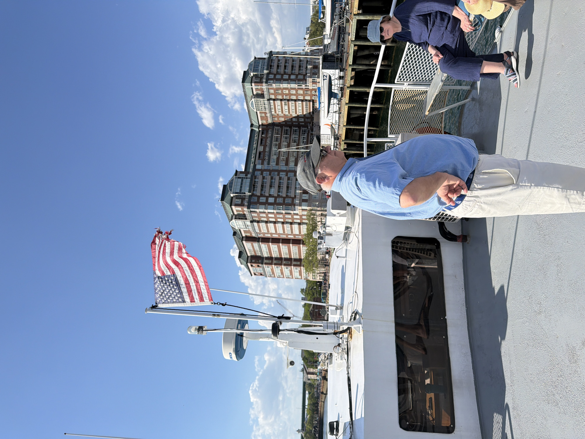 The image depicts a scene from the deck of a boat, likely in Boston, Massachusetts. A man wearing a blue shirt, khaki pants, and a hat is standing on the deck, with his back partially to the camera. To his right, another individual wearing a blue outfit and a straw hat is sitting. In the background, there are brick buildings indicative of Boston's architecture, suggesting the boat is likely traveling along the city's waterfront. An American flag is prominently displayed on a pole attached to the boat, further suggesting the location in the United States. The sky is clear and blue with scattered white clouds. The scene appears to be taken during the daytime.
