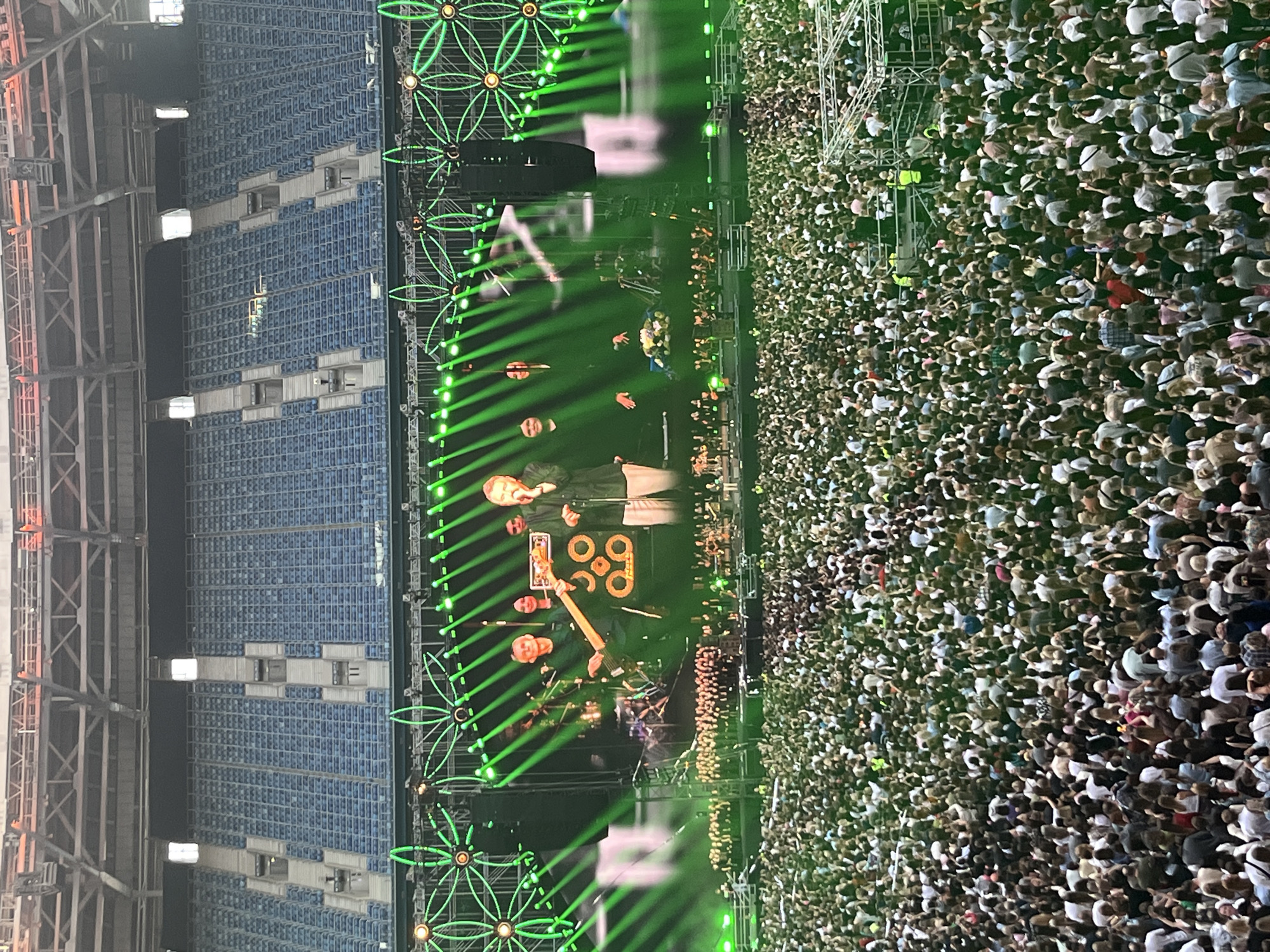 This is a high-angle, vertically oriented, wide shot of a concert taking place in an outdoor stadium. The stadium has tiered blue seating, with a large crowd of people filling the space in front of the stage. The crowd is densely packed, with many people wearing light-colored clothing, suggesting it might be daytime or evening with bright lighting.
On the stage, there are several performers, with one individual playing a guitar. Green lights are prominent, projecting beams and illuminating decorative floral-like structures. There is also a large screen or display behind the performers showing an orange graphic with the number "99". The overall atmosphere appears energetic and celebratory, as suggested by the large gathering and stage production.
The setting appears to be a large stadium in Saint Petersburg, Russia, indicated by the provided location context. The time of day is difficult to ascertain precisely due to the stage lighting, but the amount of visible light on the crowd suggests it is not deep night. The weather appears to be clear, as there are no visible signs of rain or adverse conditions. The prominent use of green lighting and the stage design contribute to a dynamic and visually striking concert experience.