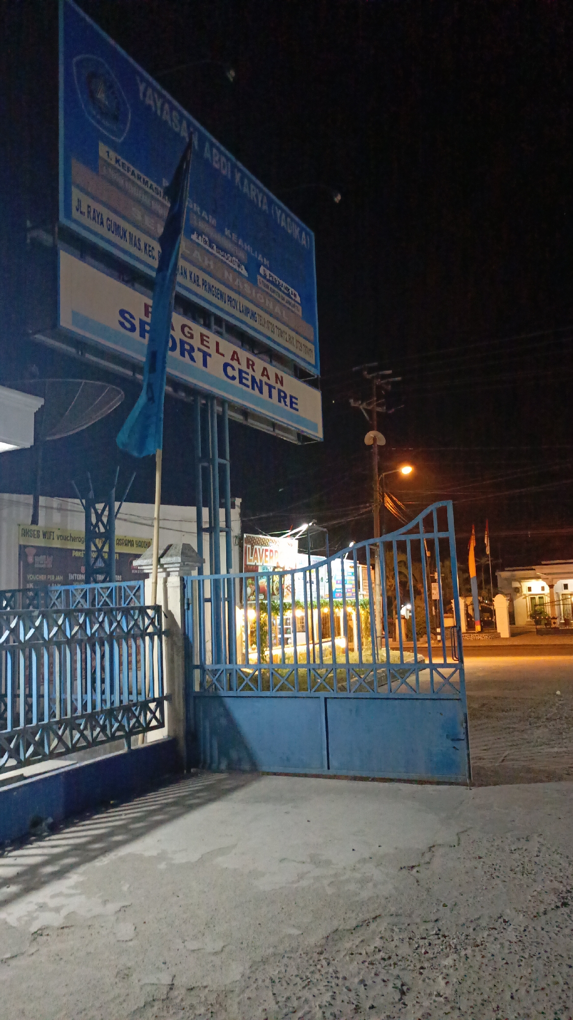The image depicts a night scene in front of the "YAYASAN ABDI KARYA (YADIKA)" or "YADIKA Foundation", located on Jl. Raya Gumuk Mas, Kecamatan Pringsewu, Kabupaten Pringsewu, Provinsi Lampung, Indonesia. A large blue billboard with white and yellow text is prominently displayed, announcing the foundation's name and various programs. Below the billboard, the text "PAGELARAN SPORT CENTRE" is visible, suggesting a sports facility.
A blue metal gate, partially open, stands in the foreground, leading into the property. The gate has an intricate criss-cross pattern on its upper section. To the left of the gate, a white building with a satellite dish is visible. Another smaller sign with text including "AKSES WIFI voucherag... ASRAMA YADIKA" can be seen. Beyond the gate, within the property, there is a building with lights illuminating it, and the word "LAVER" is discernable on a sign. Two flags, one orange and one possibly red, are planted on poles to the right of the entrance.
The environment is dark, with streetlights and lights from the buildings casting illumination on the scene. The ground in the foreground is concrete, with shadows from the gate and fence cast upon it. Electrical poles and wires are visible in the background, indicating an urban or semi-urban setting. No people are visible in the image.
