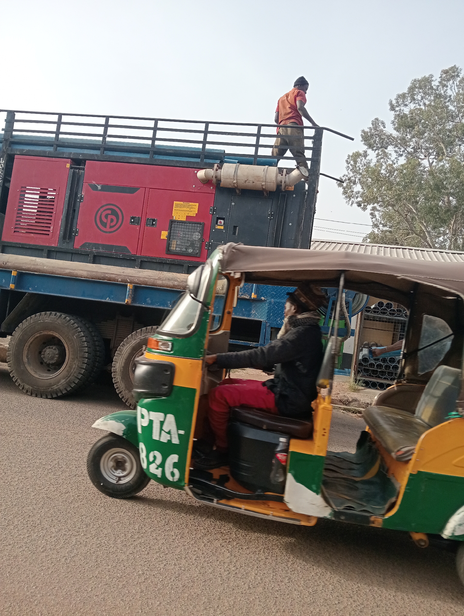 A man is standing on the back of a flatbed truck with a large, red generator. The truck is driving down a paved road in Jos, Nigeria. A yellow and green auto rickshaw ("Keke Napep") with the license plate "PTA-826" is in the foreground of the photo. The auto rickshaw driver is wearing a black jacket and red pants. A Coca-Cola bottle is visible behind the driver. The scene is taken during daylight hours. The presence of the auto rickshaw and the style of the generator truck indicate the location as Jos, Nigeria. The person atop the truck wears an orange shirt and tan pants.