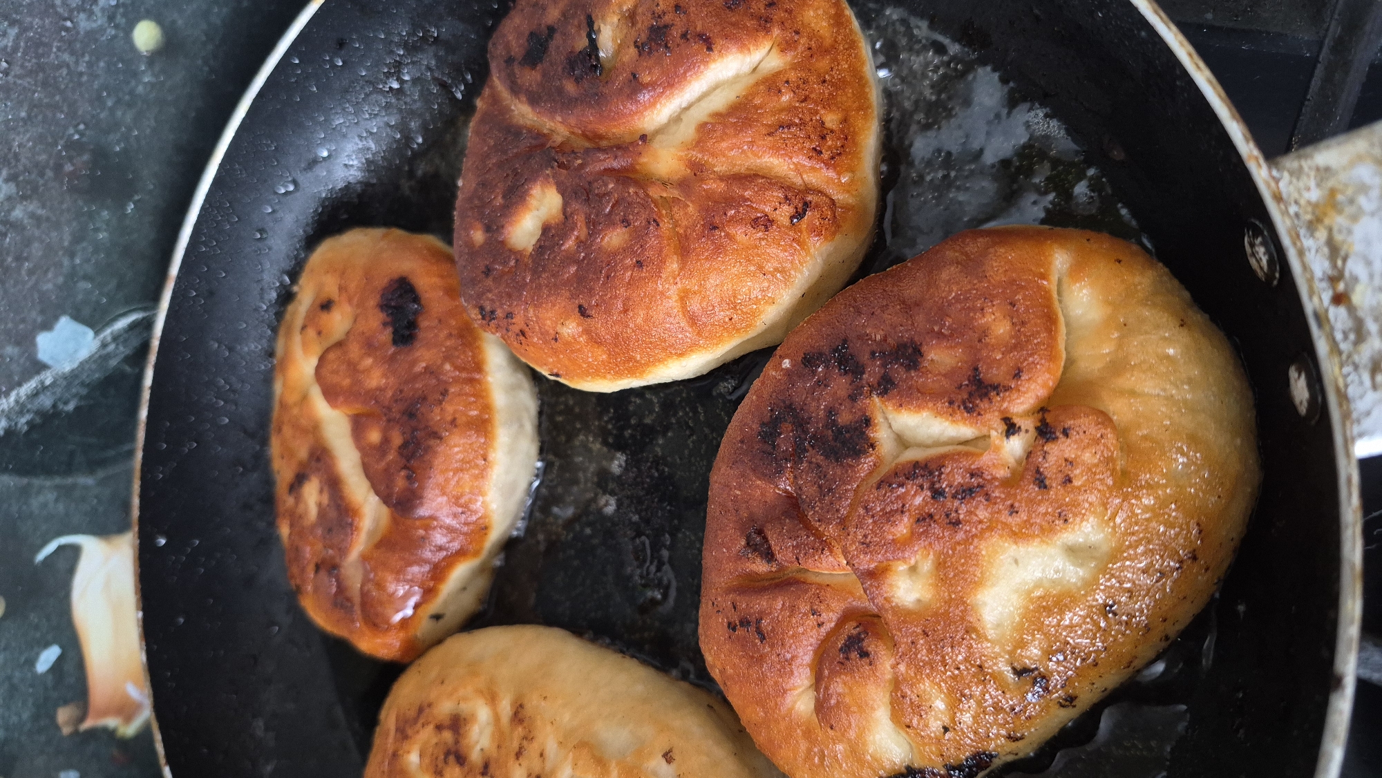 In a black frying pan, four golden-brown, round pastries are being fried in oil. The pastries have a slightly puffed appearance and some areas are darker brown, indicating a crispy texture. The pan is resting on a stovetop. The background is slightly out of focus, showing a patterned surface. There are no people visible in the image. The scene appears to be a domestic kitchen setting.
