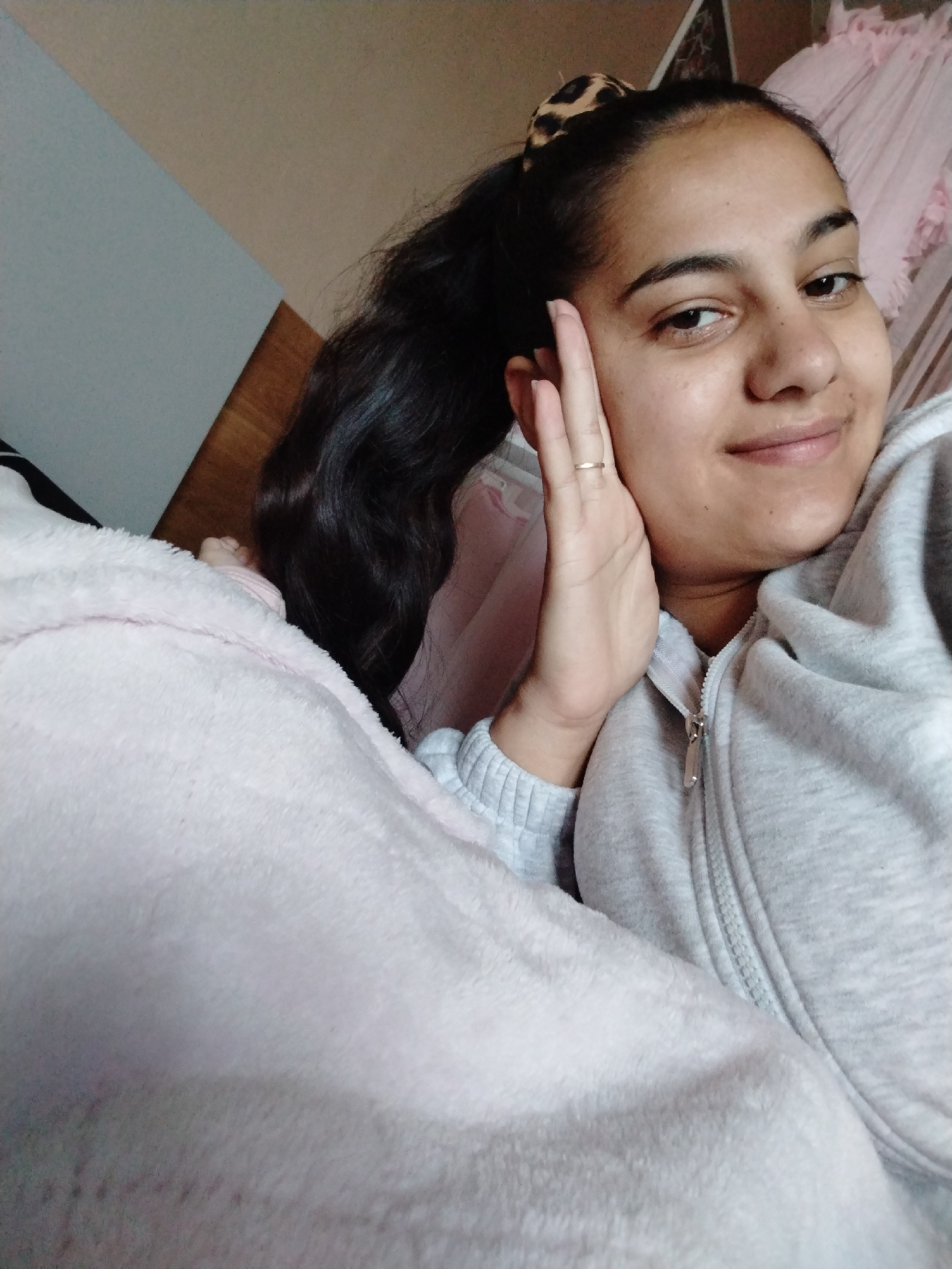 A woman with dark hair pulled back in a leopard-print scrunchie, wearing a gray zip-up hoodie, is lying under a pink blanket. She is holding her hand up to her face and smiling. The background includes a wall with two different colors, as well as pink fabric draped over something. The location is Unknown, Unknown.