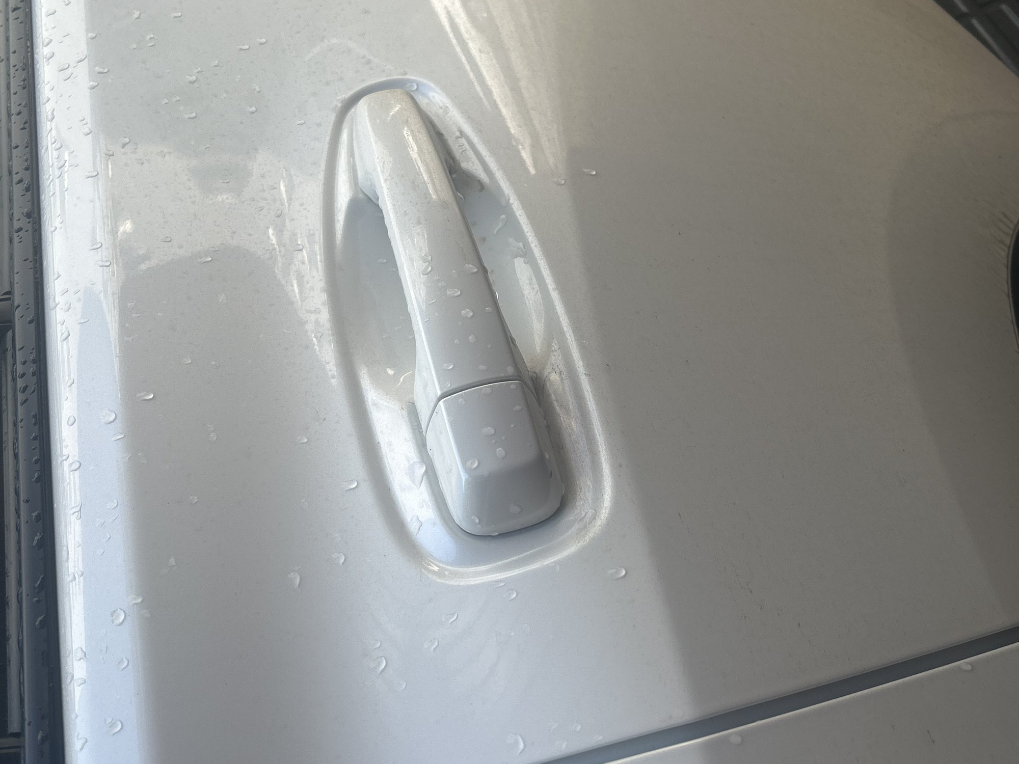 The image shows a close-up of a white car door handle with water droplets on its surface. The car appears to be parked outdoors, possibly after being washed or in rainy weather. The sunlight reflects off the wet surface, creating bright highlights. The scene is set in Perm, Russia. There is no visible text in the image. The focus is on the texture and details of the car's exterior, with the water droplets adding a sense of freshness or recent exposure to moisture. The overall mood is neutral and observational, highlighting the simple details of the car's surface.