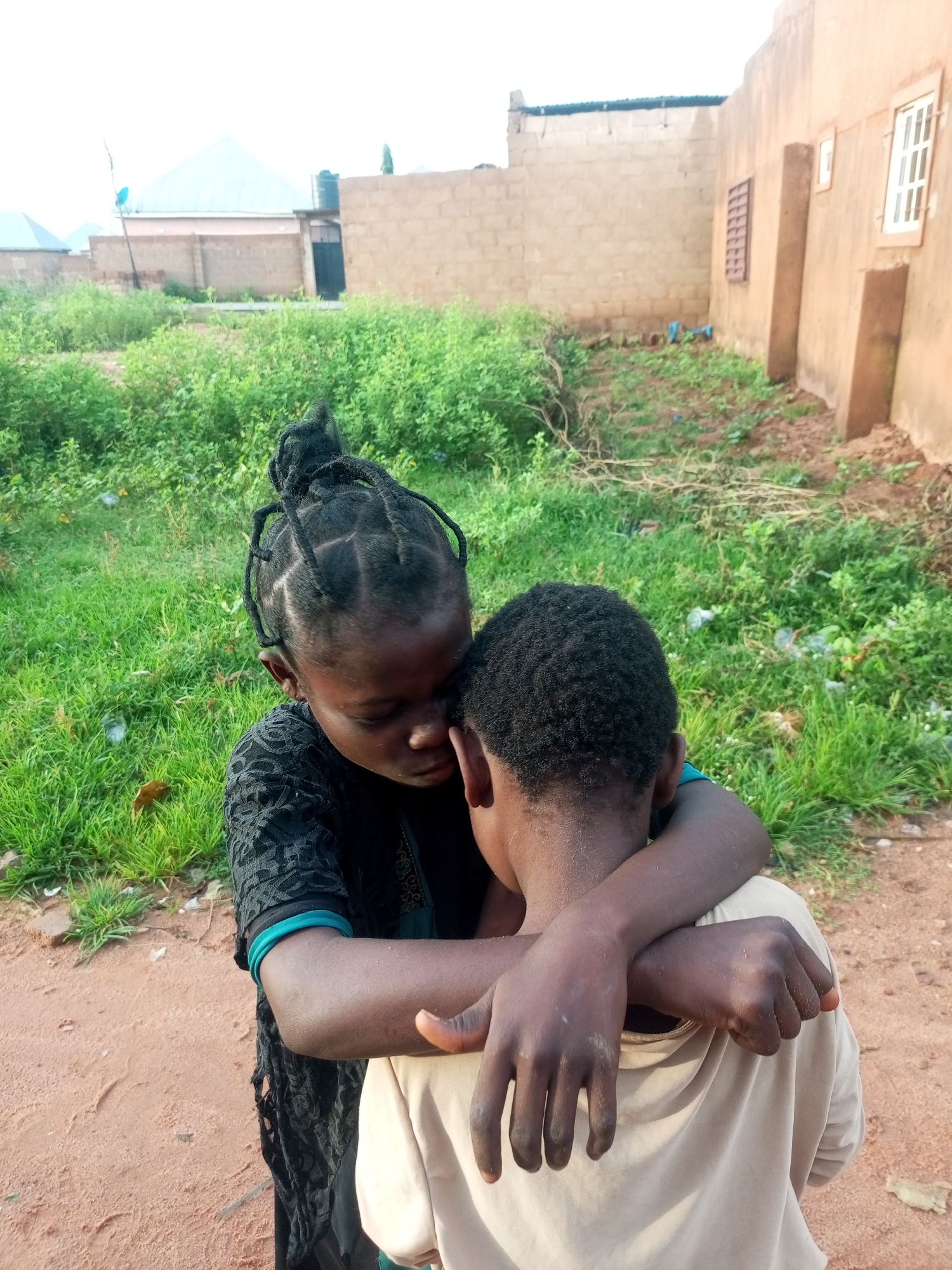 In Tempure, Nigeria, two young Black children are pictured embracing each other. The child on the left, with braided hair pulled into a bun, wears a black patterned top with turquoise trim. Their arms are wrapped around the shoulders of the child on the right, who has short, curly hair and wears a light-colored t-shirt. They are standing outdoors on a dirt path. In the background, there is overgrown green vegetation and buildings constructed from tan brick. One building has a visible window with white panes and another has a barred window. The sky is overcast.