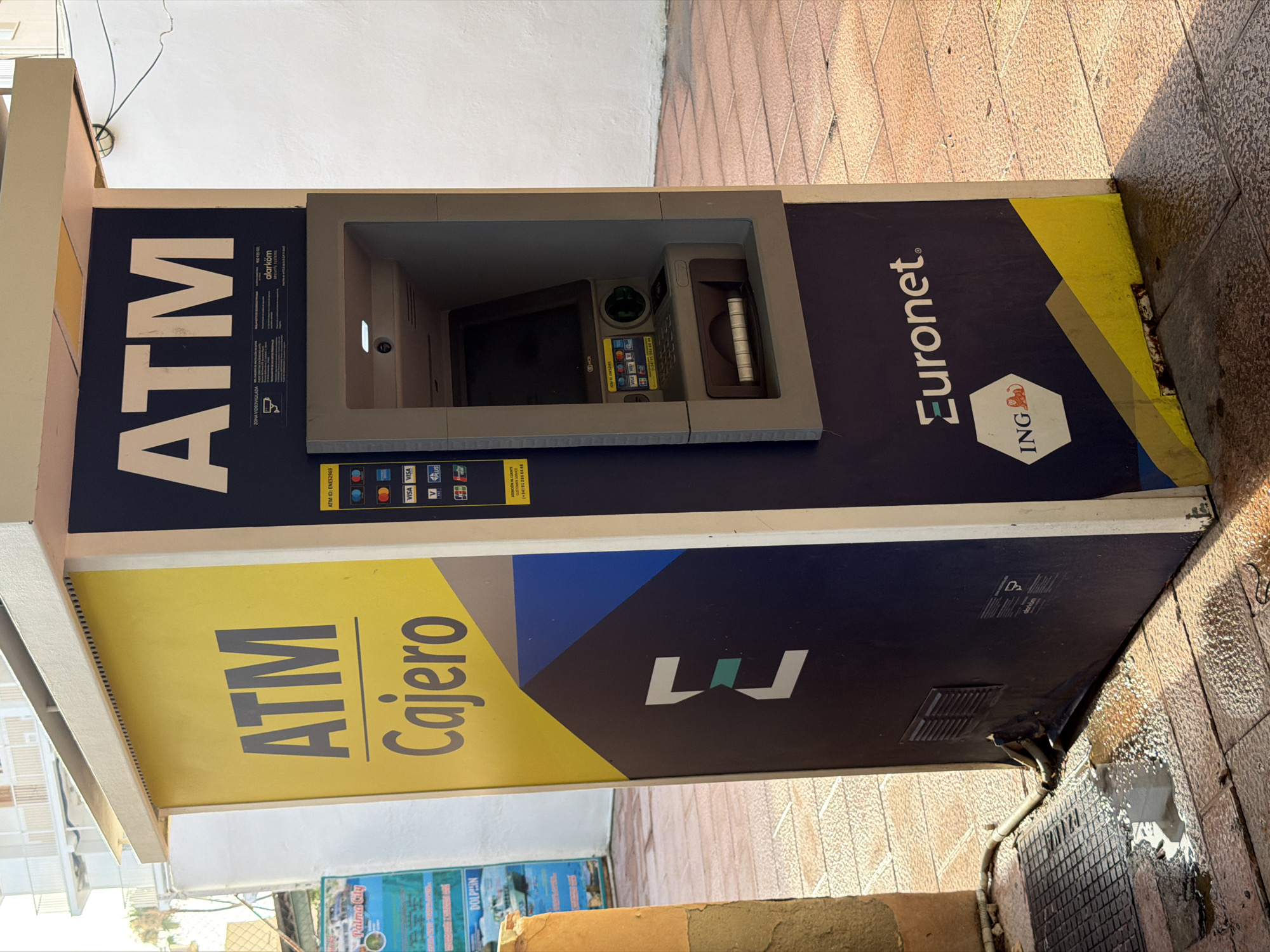 An outdoor ATM machine, branded with "Euronet" and "ING," is situated on a tiled pathway in Magaluf, Spain. The ATM is housed within a yellow and dark blue structure featuring large "ATM" lettering on its left side, and the "Euronet" logo prominently displayed on its front. The machine itself has a screen, keypad, card slot, and cash dispenser. Next to the ATM, a metal grate is visible on the ground, with some water pooling around it and near the base of the ATM. The background shows a building with balconies and a white wall. The lighting suggests it is daytime, with shadows cast by the structure.