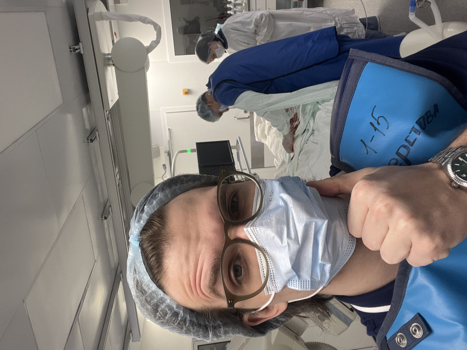 A male medical professional in a sterile environment, likely a catheterization lab or operating room in Moscow, Russia, takes a selfie. He wears a light blue disposable hairnet, brown-rimmed eyeglasses, and a light blue surgical mask. His attire includes a dark blue scrub top and a light blue lead apron, which has "1-15" and "СЕРВЕТКА" (Cyrillic for "napkin," or more likely a functional code/label) written in black marker. His right hand is positioned near his chin, potentially adjusting his mask or apron, revealing a silver wristwatch on his left wrist.
In the midground, two additional medical personnel are engaged in a procedure. One individual, wearing a blue scrub top, hairnet, and lead apron, leans over an operating table where a patient is draped in white and light green sterile sheets. Red staining, consistent with blood, is visible on the drapes. This individual wears white gloves, indicating active involvement in the procedure. The second person, in a white gown and hairnet, stands further to the right near a counter with various medical supplies.
The background reveals a brightly lit, clinical room with white walls and a white drop ceiling. Visible medical equipment includes a large imaging system (possibly a C-arm) on the left, a monitor displaying a green light, and overhead spherical light fixtures.