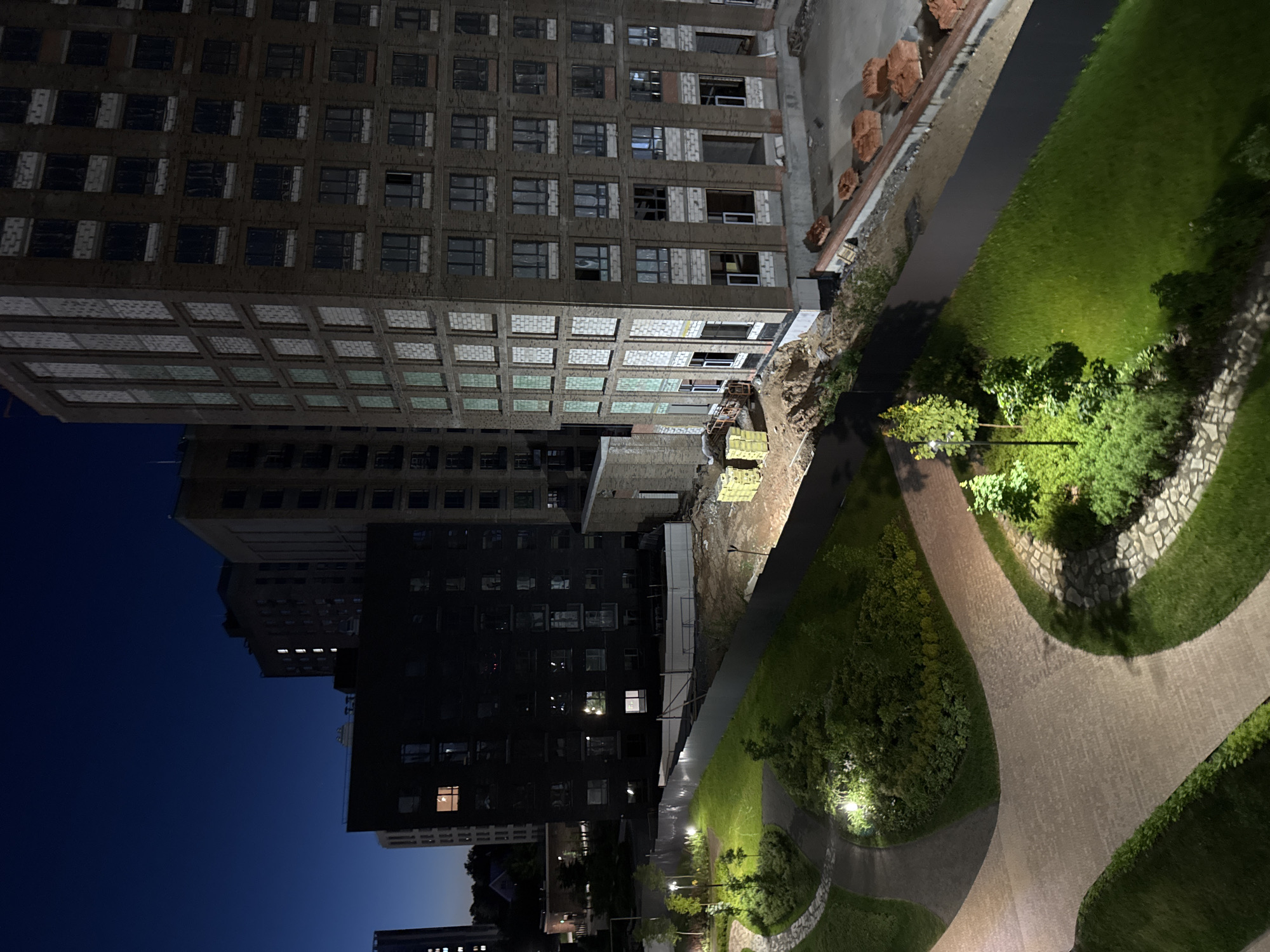 The media file captures an urban landscape at night or deep twilight, likely from a high vantage point overlooking buildings, a construction site, and a landscaped park in Izhevsk, Russia. The sky is a deep, dark blue, suggesting late evening or early morning hours.
In the mid-ground, a large, multi-story building, possibly under construction or renovation, dominates the scene. Its facade appears to be a brick-like material, featuring numerous windows, some open frames, others covered, indicating different stages of completion. The lower floors of this building exhibit a distinct grid-patterned facade. Adjacent to this structure is a construction area with piles of reddish bricks, exposed earth, and stacks of yellow building materials.
Behind the prominent building, another tall, dark structure is visible, illuminated by several glowing windows. Further in the distance, other buildings are silhouetted against the dark sky, marked by scattered lights.
In the foreground and mid-ground, a well-maintained park or courtyard offers a contrast to the surrounding architecture. It features lush green grass, illuminated by various spotlights, and winding paved pathways that curve through the landscape. These paths are possibly made of cobblestone or patterned concrete. Numerous trees and bushes are planted throughout the park, casting shadows under the artificial lighting from pathway lights and spotlights. Decorative stone retaining walls or borders are also visible along the paths.
No people or specific activities are depicted in the static scene. The only implied activity is the ongoing construction. There is no visible text in the image. The scene conveys a sense of quiet urban development observed in the tranquil hours of the night.