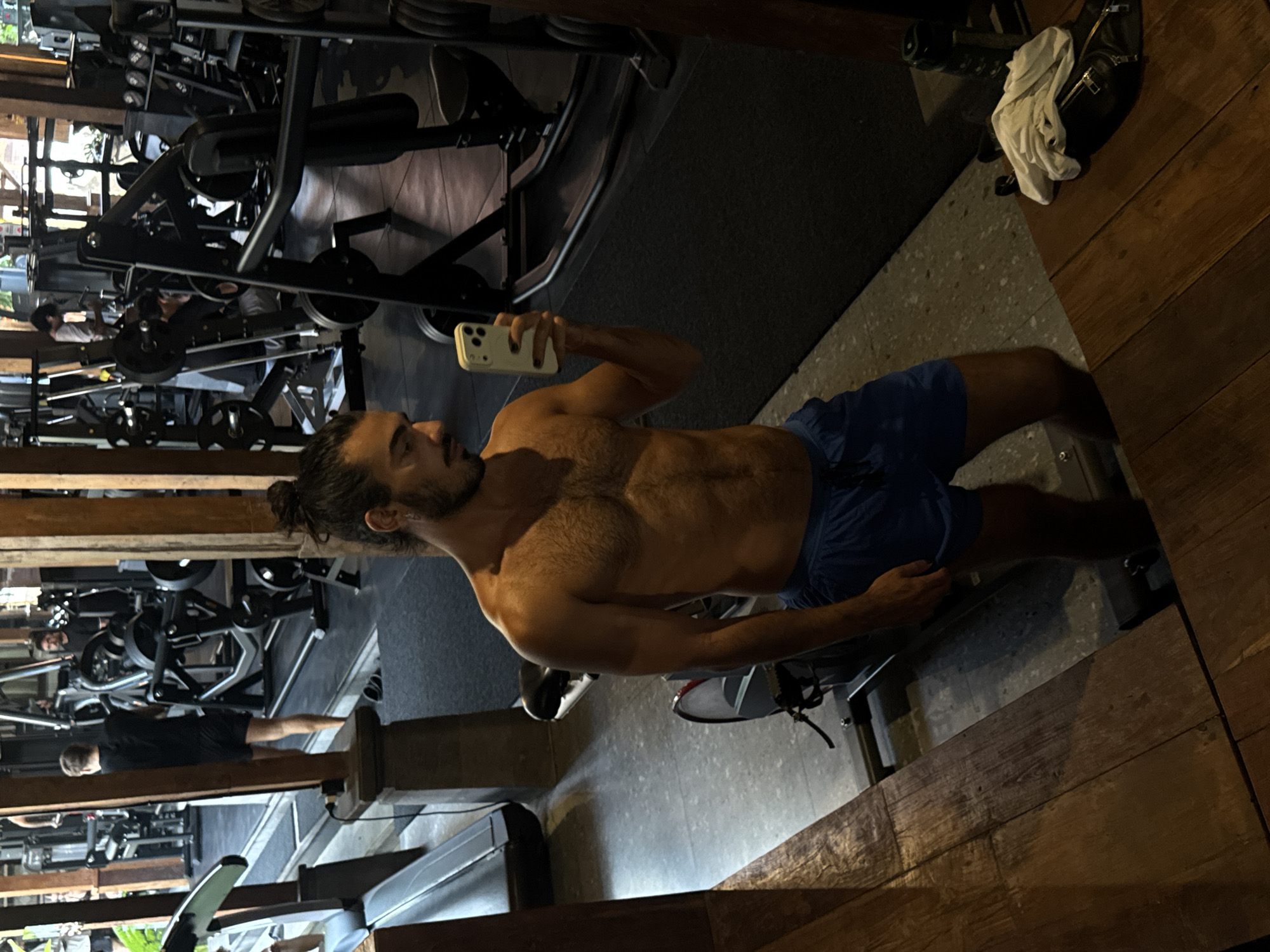 In a gym in North Kuta, Indonesia, a shirtless man with dark, curly hair tied back in a bun and a beard is taking a mirror selfie. He is wearing blue athletic shorts. He is standing next to a stationary exercise bike and is holding a white smartphone in his right hand, capturing his reflection. The gym features various weight machines and equipment, with wooden accents and a stone floor. In the background, other gym-goers are visible, engaged in their workouts. A treadmill is partially visible on the left, and a wooden bench with a towel and a black fanny pack is on the right.