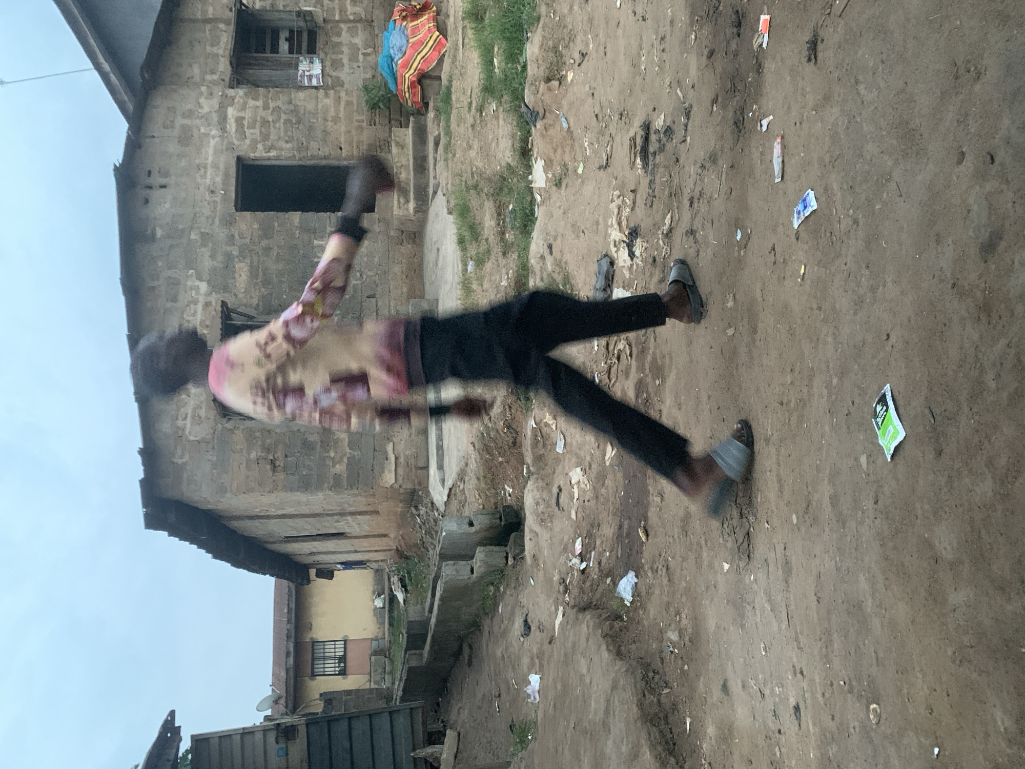 In Shagamu, Nigeria, a young Black person is captured mid-stride in an outdoor scene. The individual, wearing a multi-colored long-sleeved shirt and dark trousers with gray slides, is moving from left to right. The background features a rustic, unpaved street littered with debris, including various pieces of white and colored paper packaging. Two low-rise buildings are visible: one constructed from rough, light-colored bricks with a corrugated metal roof and an open doorway, and another to its left, painted yellow with a barred window and a more modern metal-clad structure adjacent. The environment suggests a less developed urban area, with evidence of disarray and sparse vegetation growing between concrete structures and along a drainage channel. The sky above is overcast.