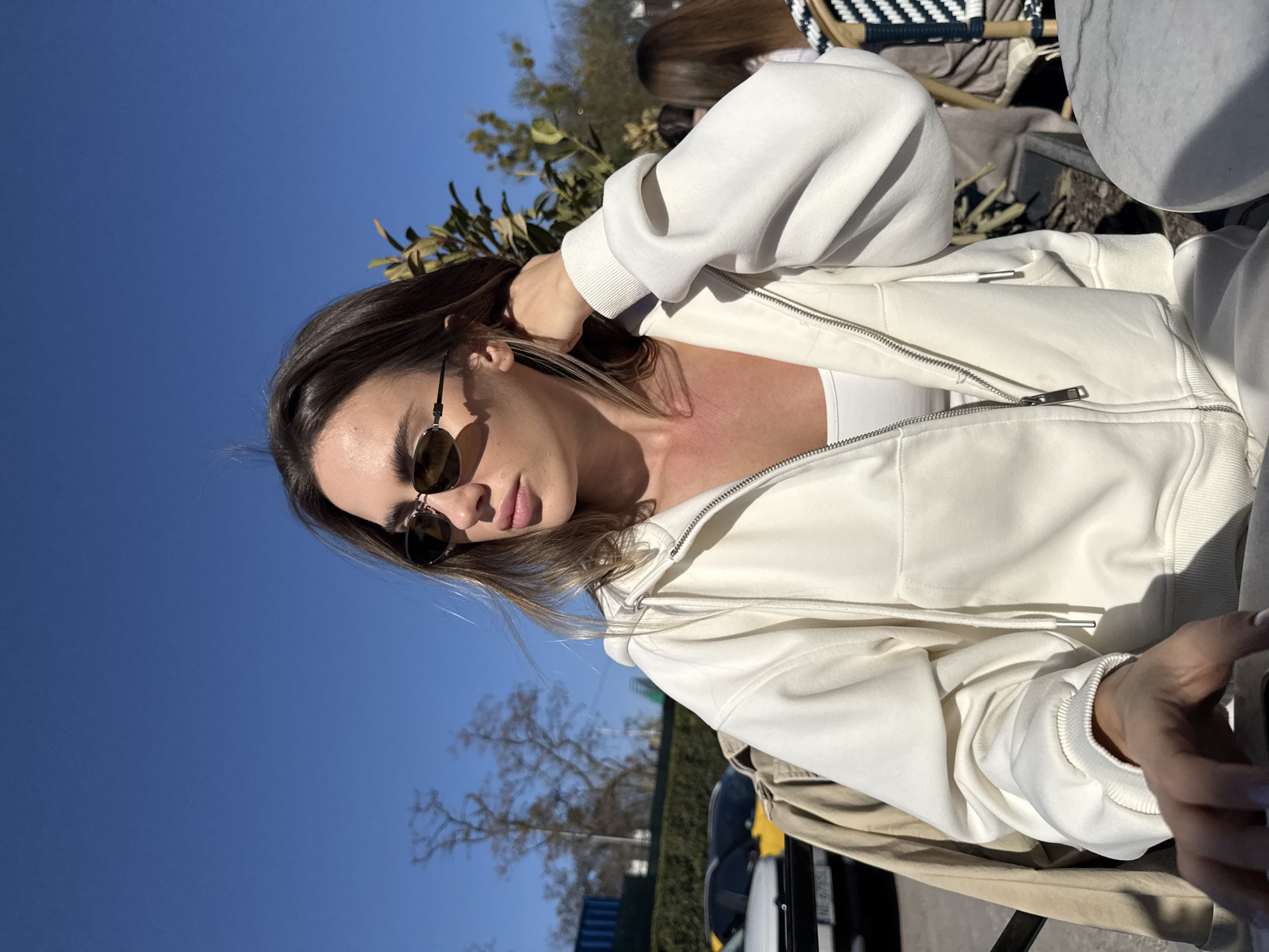 The image features a young woman captured in an outdoor setting on a clear, sunny day, likely in Warsaw, Poland. She is positioned with her head tilted, looking down and to her right, wearing dark-tinted sunglasses that obscure part of her face. Her right hand is raised, gently touching the back of her head or hair, while her left arm rests, possibly holding a mobile device.
She is dressed in a cream or off-white hooded zippered jacket, worn over a white top. The jacket has ribbed cuffs and a silver-colored zipper. Her skin, particularly on her chest and neck, shows a light flush or tan.
The background reveals a vibrant, cloudless blue sky dominating the upper half of the frame. Scattered tree branches, some with green foliage, are visible against the sky. Further back, blurry elements suggest an urban environment, including what appears to be the roof of a car and possibly other structures. Another person's head and shoulder are faintly visible behind her to the right, suggesting she is not alone. A distant license plate shows partial text, possibly "NJ-672-SD". The bright sunlight and absence of harsh shadows indicate either mid-morning or late afternoon. The overall mood is relaxed and peaceful.
