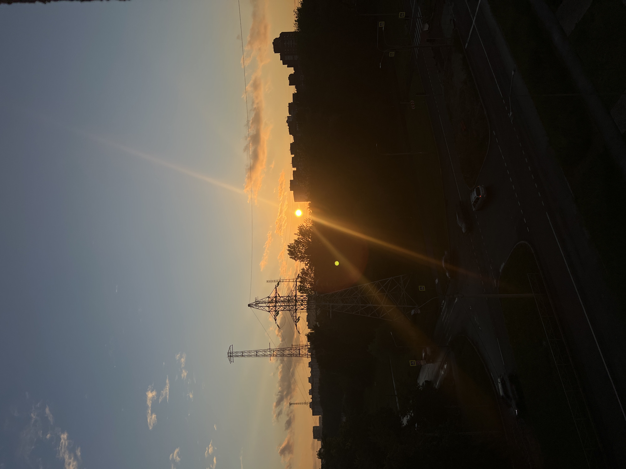This is a vertical, low-angle photograph taken at sunset in Saint Petersburg, Russia. The scene captures a cityscape silhouetted against the warm hues of the setting sun. In the foreground, two large power line towers dominate the frame, their skeletal structures stark against the sky. Beyond them, a line of apartment buildings forms a dark, jagged horizon. The sky is a gradient of blues and oranges, with wispy clouds catching the last rays of sunlight.
To the right of the frame, a road curves into the distance, its asphalt dark and reflecting some of the ambient light. A few streetlights are visible along the road. A white car is parked on the side of the road, its brake lights faintly visible. The overall impression is one of an urban environment at the end of the day, with the natural beauty of the sunset contrasting with the man-made structures. The low angle emphasizes the scale of the power towers and the buildings, giving the scene a somewhat imposing feel. The time of day is clearly sunset, with the sun low on the horizon and casting long shadows. The weather appears clear, with the sun breaking through the clouds. No text is visible in the image.