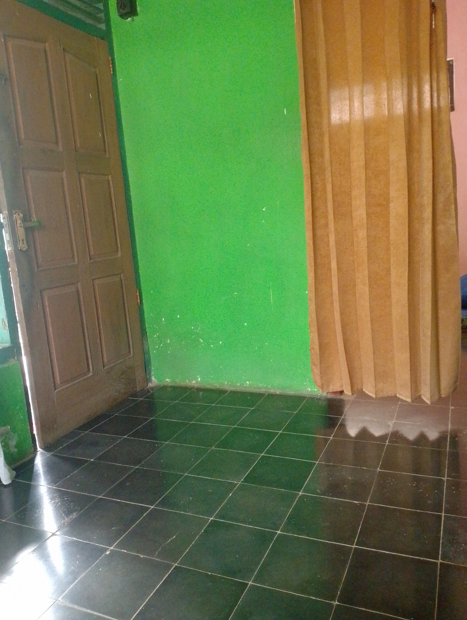 The image depicts an interior scene in Sukamanah, Indonesia, focusing on a corner where a brown wooden door meets a bright green wall. The door, slightly ajar, reveals a sliver of a lighter-colored interior and has a silver and brass door handle. To the right of the green wall, a light brown pleated curtain hangs, obscuring whatever lies beyond. The floor is tiled with large, dark, square tiles that are highly reflective, showing streaks of light and the faint outlines of the pleated curtain. Some small white speckles are visible on the green wall and on the floor tiles. The lighting suggests it is daytime, with natural light entering from the left, casting a bright glare on the floor near the door. The scene is static and there is no visible text. The overall impression is of a simple, possibly residential, interior space.