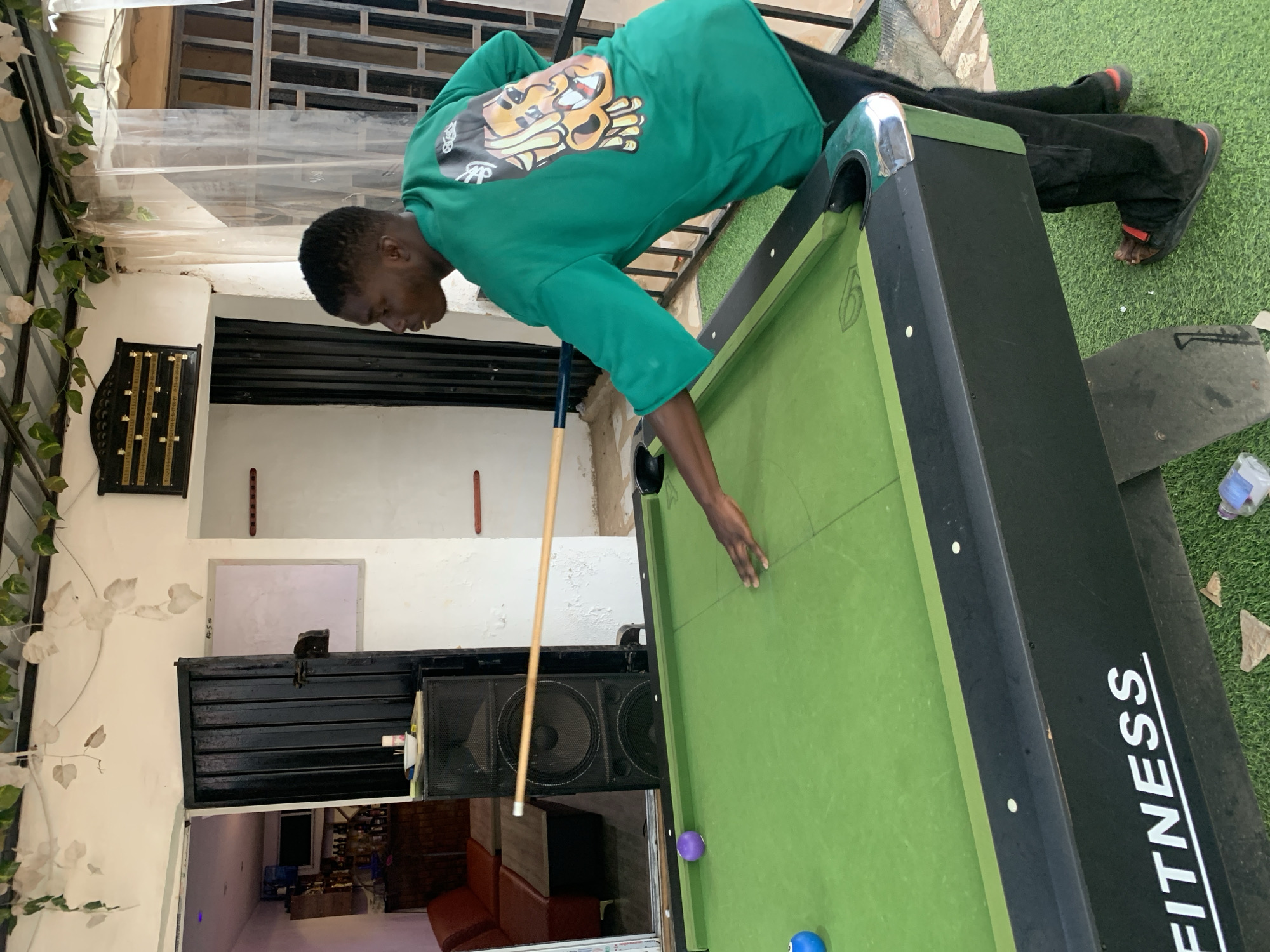 A man wearing a green long-sleeved shirt with a cartoon graphic, black pants, and sandals plays pool on a table with green felt in an outdoor setting in Barakin Akawo, Nigeria. He leans over the table with his left hand on the surface and holds a pool cue in his right hand, poised to strike a ball. The table has black trim and the word "FITNESS" printed vertically on one side. The background includes a building with a window, a speaker system, and a glimpse of an interior room with a red couch. A section of artificial turf is visible on the ground near the pool table.