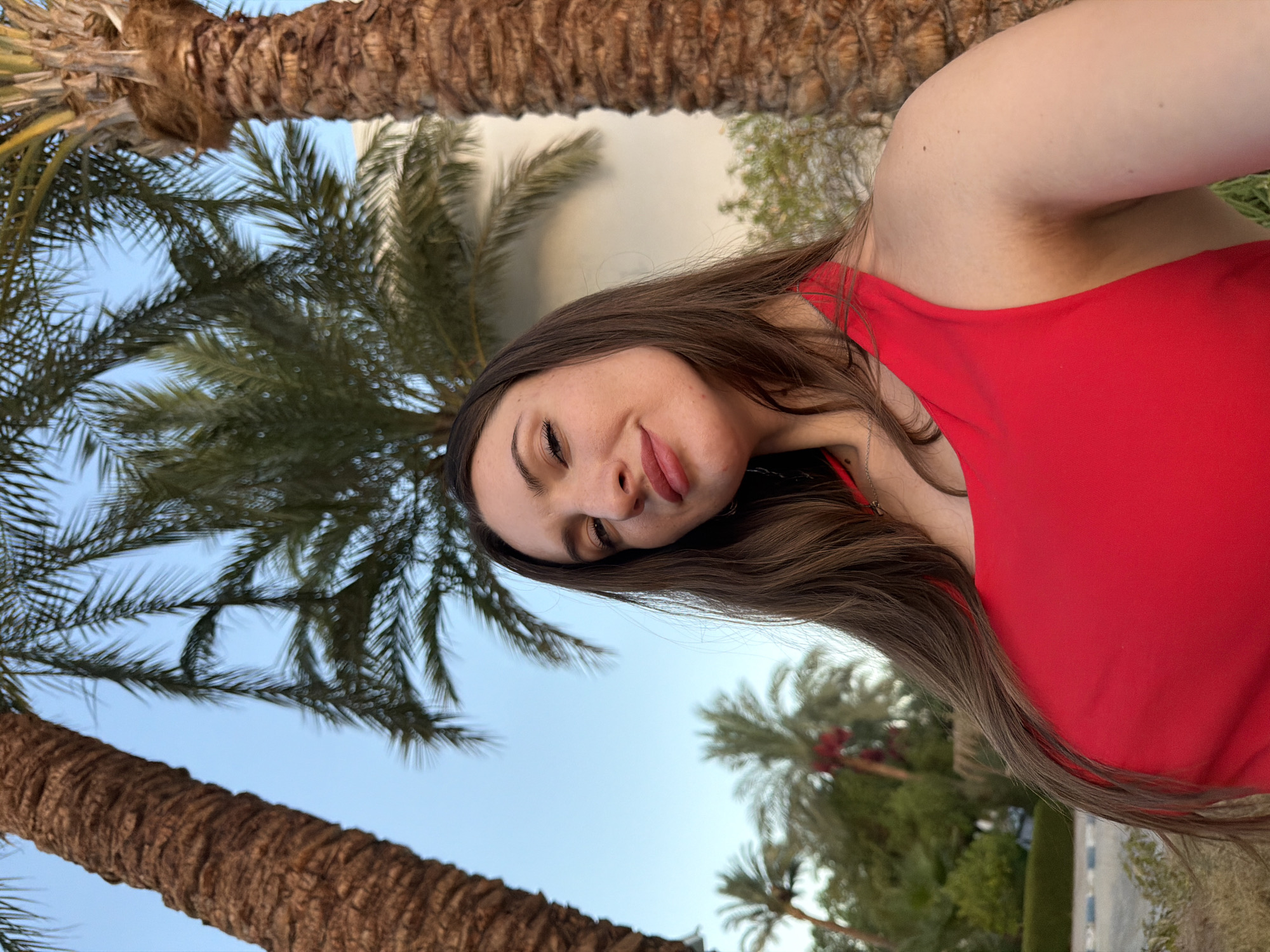 The image shows a woman in a red swimsuit posing for a selfie outdoors. She has long brown hair and is looking towards the camera with a slight smile. She is framed by two large palm trees, and other palm trees and greenery are visible in the background, along with a building and the sky. The overall setting suggests a tropical or resort location. The woman is the central subject of the image.