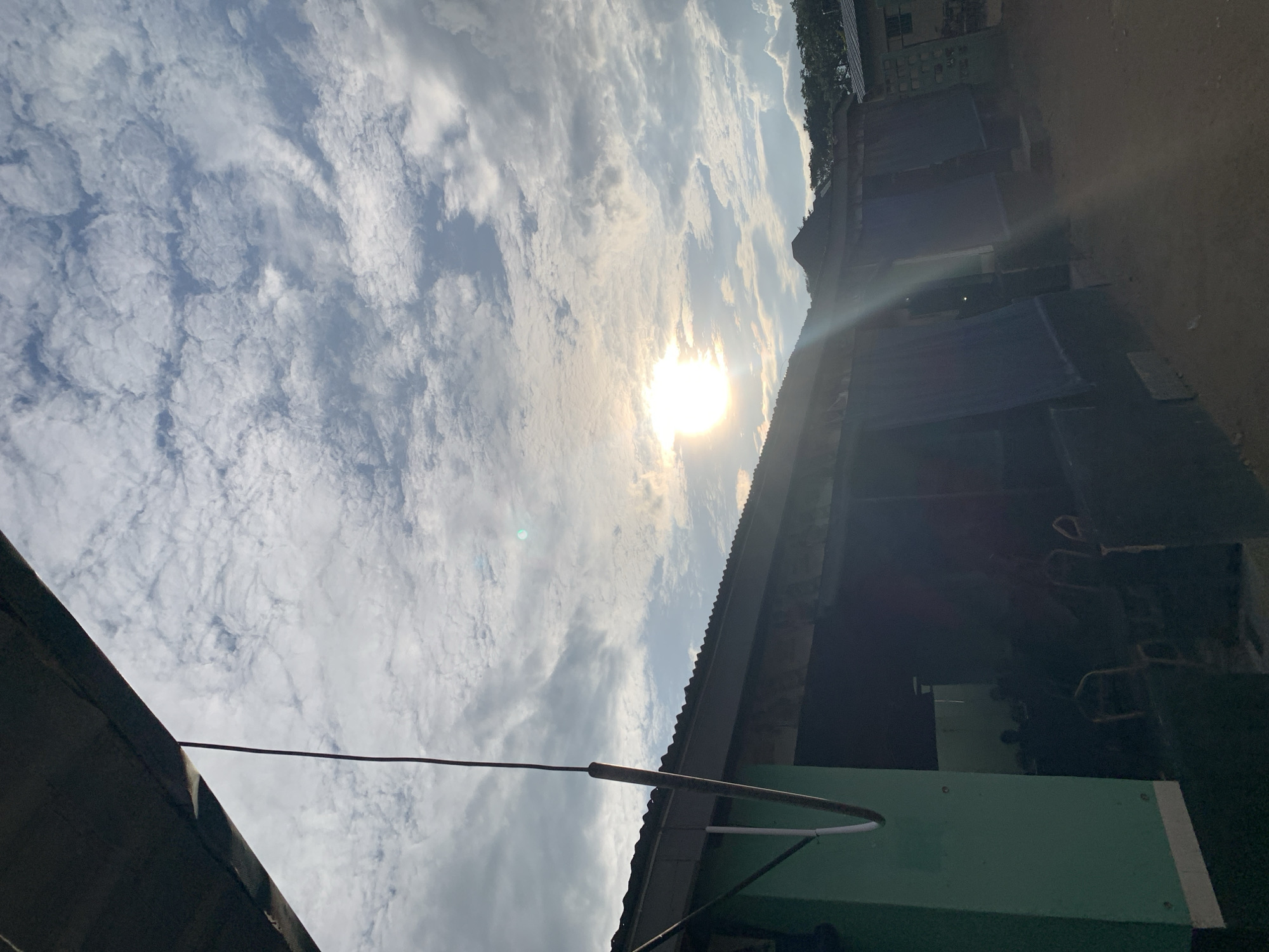 This image captures an outdoor scene in the city of New Stadium, Nigeria, under a partly cloudy sky with prominent sunlight. The upper portion of the frame is dominated by a bright, diffused sunburst located in the upper-middle right, casting significant lens flare downwards and to the right. The sky exhibits a mix of white altocumulus and stratocumulus clouds interspersed with patches of light blue sky.
In the upper left quadrant, the underside of a dark, angled roof or overhang is visible, extending diagonally into the frame. A dark, thin cable or pipe hangs vertically from this structure, extending downwards.
On the left side of the midground, a substantial, vertically oriented structure painted in a light green hue is present, exhibiting white trim along its base and what appears to be a white pipe or conduit attached to its surface.
The central and right midground feature a series of low-rise buildings with corrugated metal roofs. The facades of these buildings are predominantly dark, with multiple rectangular openings obscured by dark, draped tarps or curtains, suggesting an attempt to block light or provide privacy. Through one of these openings on the left-most building, faint silhouettes of multiple individuals seated on chairs are discernible, indicating an occupied interior space.
The ground directly in front of these structures is unpaved, appearing as compacted dirt or earth with varying light and shadow patterns. In the background, beyond the buildings, a dense expanse of trees and foliage forms a hillside or forested area against the horizon. To the far right, a portion of another building with what appears to be a textured wall is partially visible. The overall lighting suggests strong daylight, creating significant contrast between brightly lit areas and deep shadows.