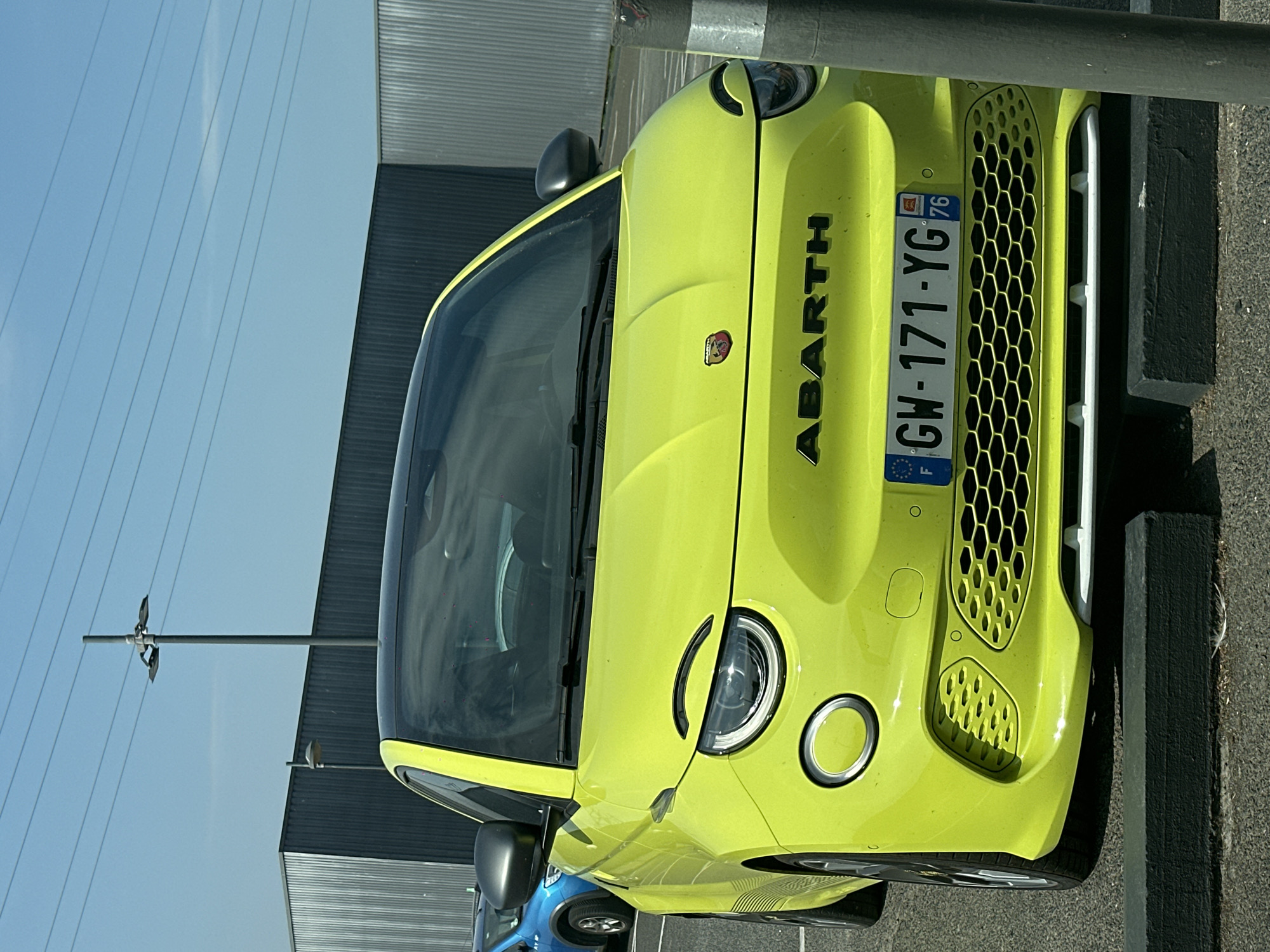 A bright yellow, compact Abarth car is parked in an outdoor lot. The car has a black roof and black side mirrors. Its headlights are rounded, giving the front of the car a somewhat personified appearance. The car's license plate reads "GW-171-YG" with French markings. In the background, a dark gray corrugated metal building is visible. A dark gray lamppost with multiple light fixtures stands in the middle ground. The scene appears to be a sunny day, as indicated by the bright lighting and clear blue sky. The car is positioned in front of a black concrete parking bumper. There are no people visible in the image. The image was taken in Mers-les-Bains, France.