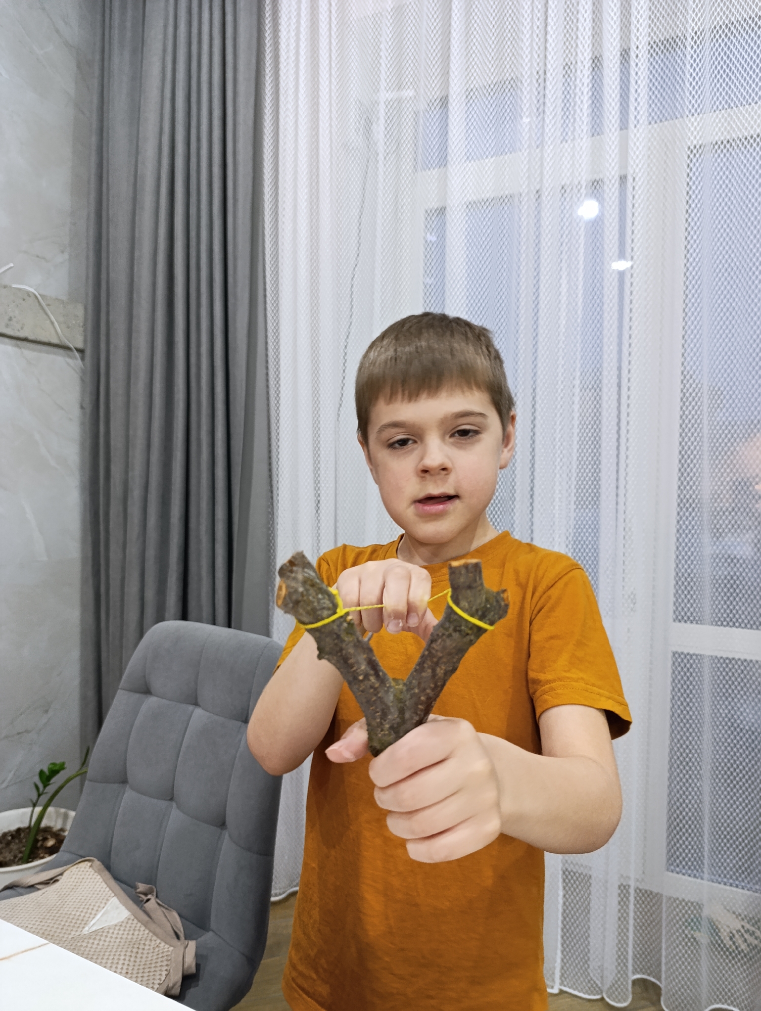 A young boy, approximately 7-10 years old with short brown hair, stands centered in the frame, indoors in what appears to be a home in Chaikovsky, Russia. He is wearing an orange short-sleeved t-shirt.
The boy is holding a homemade-looking slingshot, crafted from a V-shaped tree branch with a bright yellow rubber band stretched across its two prongs. His right hand is positioned to pull back the rubber band, while his left hand grips the base of the wooden frame. His expression is focused and slightly open-mouthed, suggesting concentration or excitement about the object.
In the background, to the left, a gray upholstered chair with a tufted backrest is partially visible, with a light-colored, patterned cloth draped over its seat. Behind the chair, a green potted plant in a white pot is present. The far background features light-colored walls, with a combination of dark gray pleated curtains on the left and sheer white, textured curtains covering a window on the right. Light filters through these curtains, indicating it is daytime. A white table surface is visible in the bottom left foreground. No visible text is present in the image.