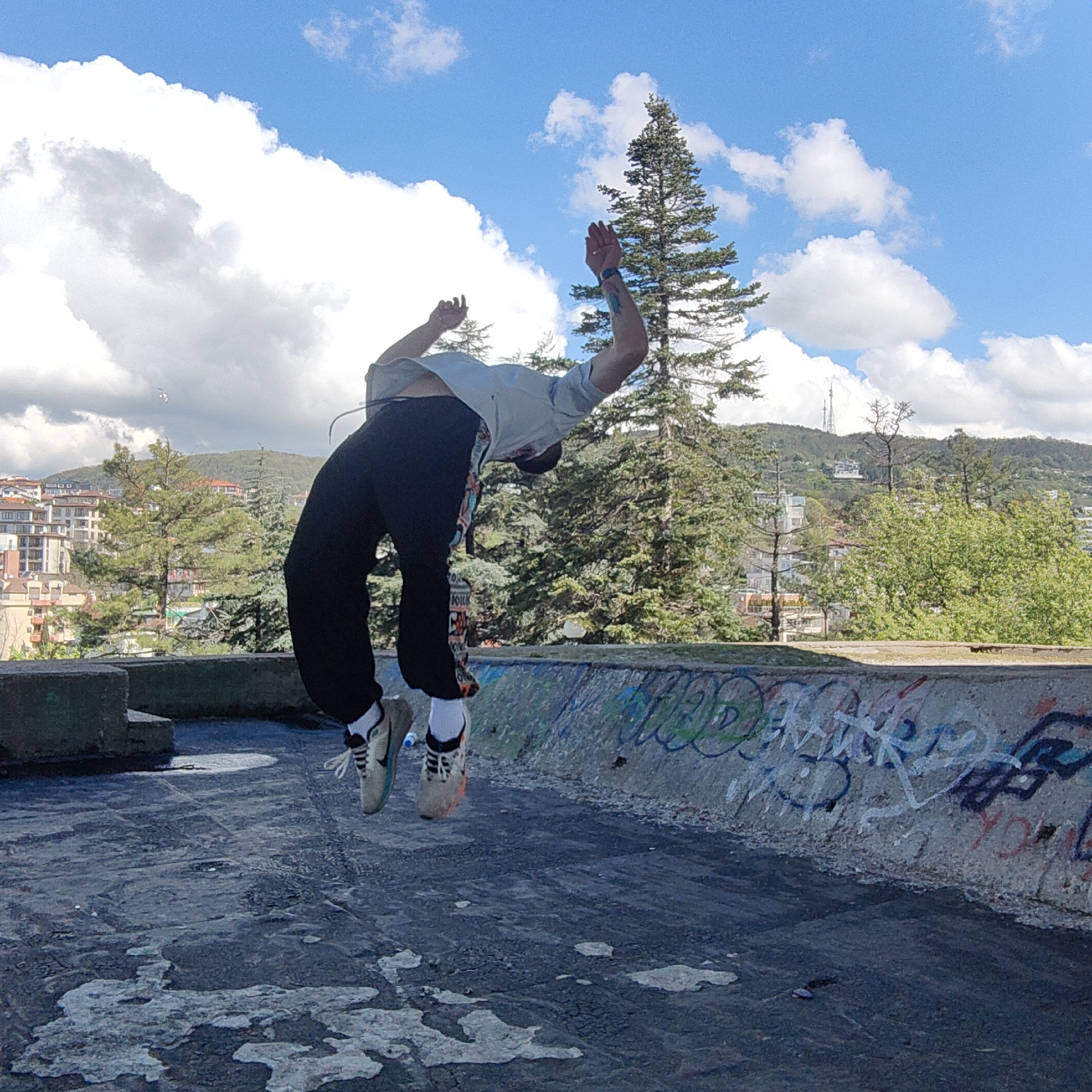 A person is in mid-air, performing a backflip on a flat, weathered concrete surface with visible graffiti along a low wall. The individual wears a light-colored shirt, dark pants, and sneakers with white socks. The background features a landscape with trees, buildings, and a hill with communication towers, under a partly cloudy sky. A white bird is visible in the sky near the buildings. The image appears to be taken in a city environment with surrounding natural elements.