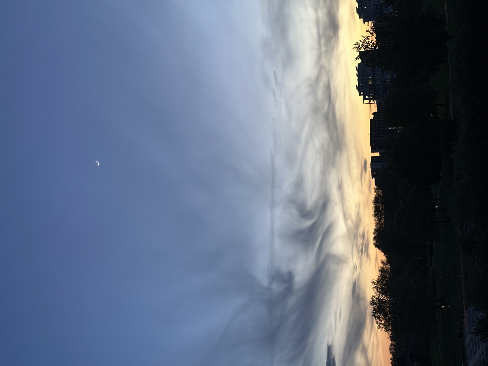 The image depicts a sky at dusk, with a crescent moon visible in the upper left portion. The sky transitions from blue to a mixture of yellow and orange near the horizon. Clouds stretch across the sky in a vertical orientation, some with a swirling appearance. Below the horizon line are buildings and trees, silhouetted against the fading light. Based on the presence of specific structures and the layout, the image likely originates from Strasbourg, France. However, without further landmarks, exact location identification remains challenging.
