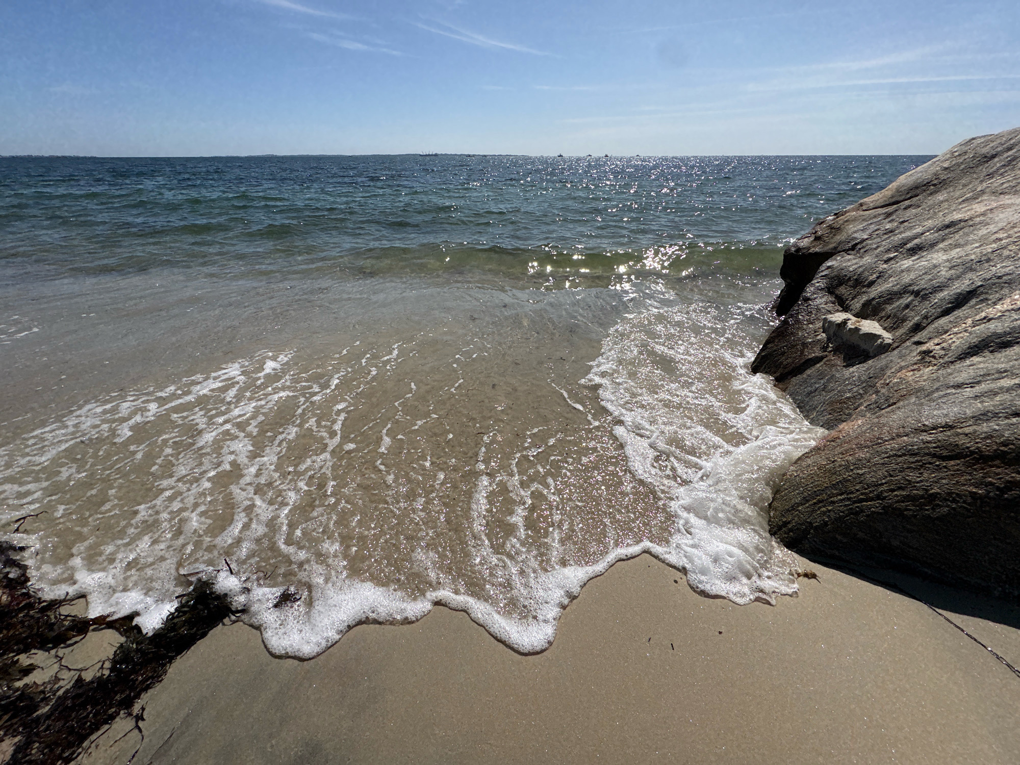 A wide-angle, eye-level shot captures a serene coastal scene on a bright, sunny day. The foreground is dominated by wet, tan-colored sand, with dark seaweed tangled at the lower left corner. A large, textured grey boulder juts into the frame from the right, its surface showing layers and veins. Gentle waves are washing onto the shore, creating foamy white water that recedes across the sand. In the midground, the clear, blue ocean stretches out, its surface sparkling with sunlight and showing gentle ripples. A distant, hazy landmass or a line of boats is visible on the horizon. The sky above is a pale blue with a few wispy white clouds. The scene is devoid of any people or animals, presenting a tranquil natural landscape. The location is identified as Waterford, United States of America, suggesting a view of the coastline there.